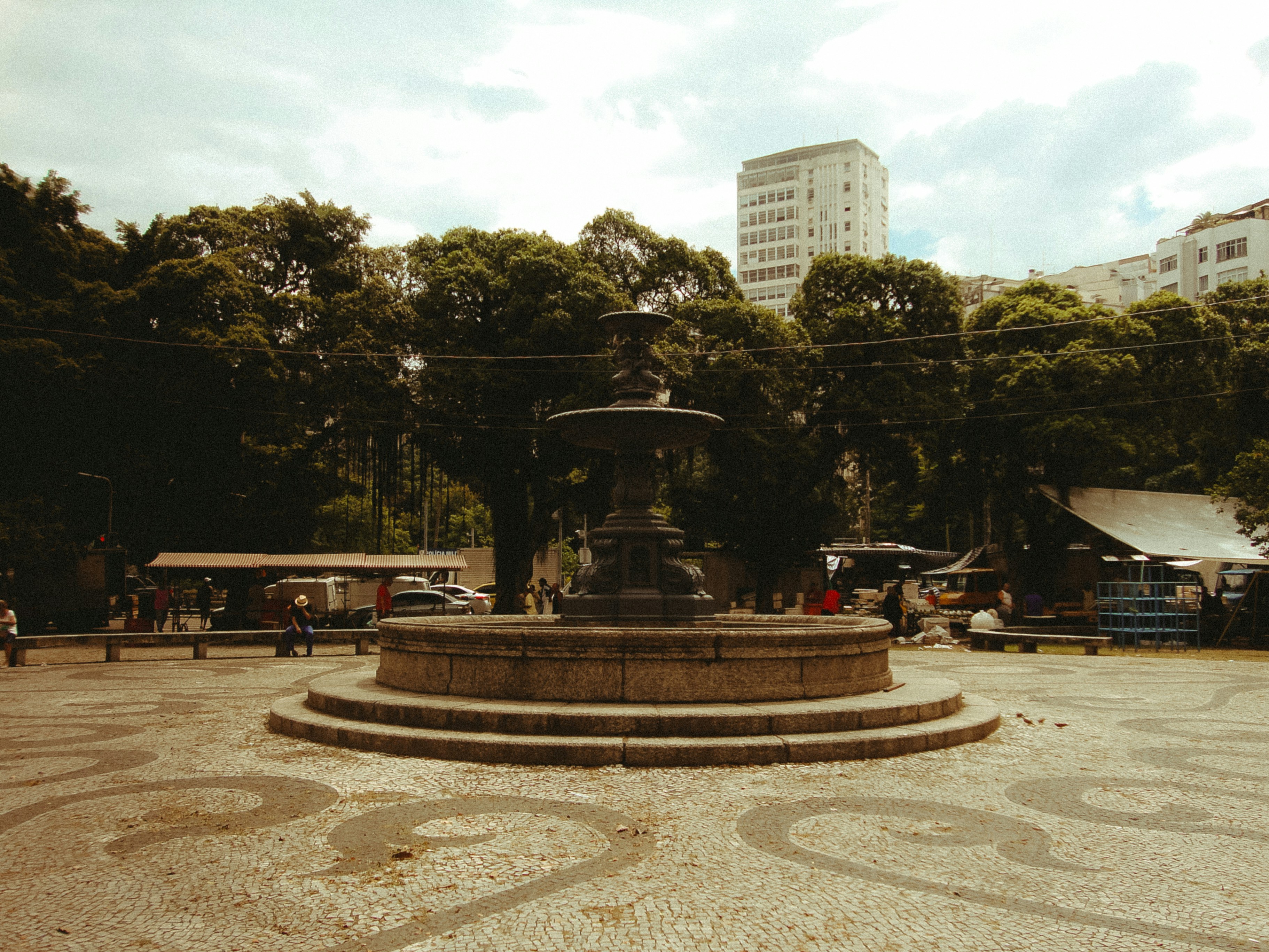 A stone fountain in a park with trees and buildings.