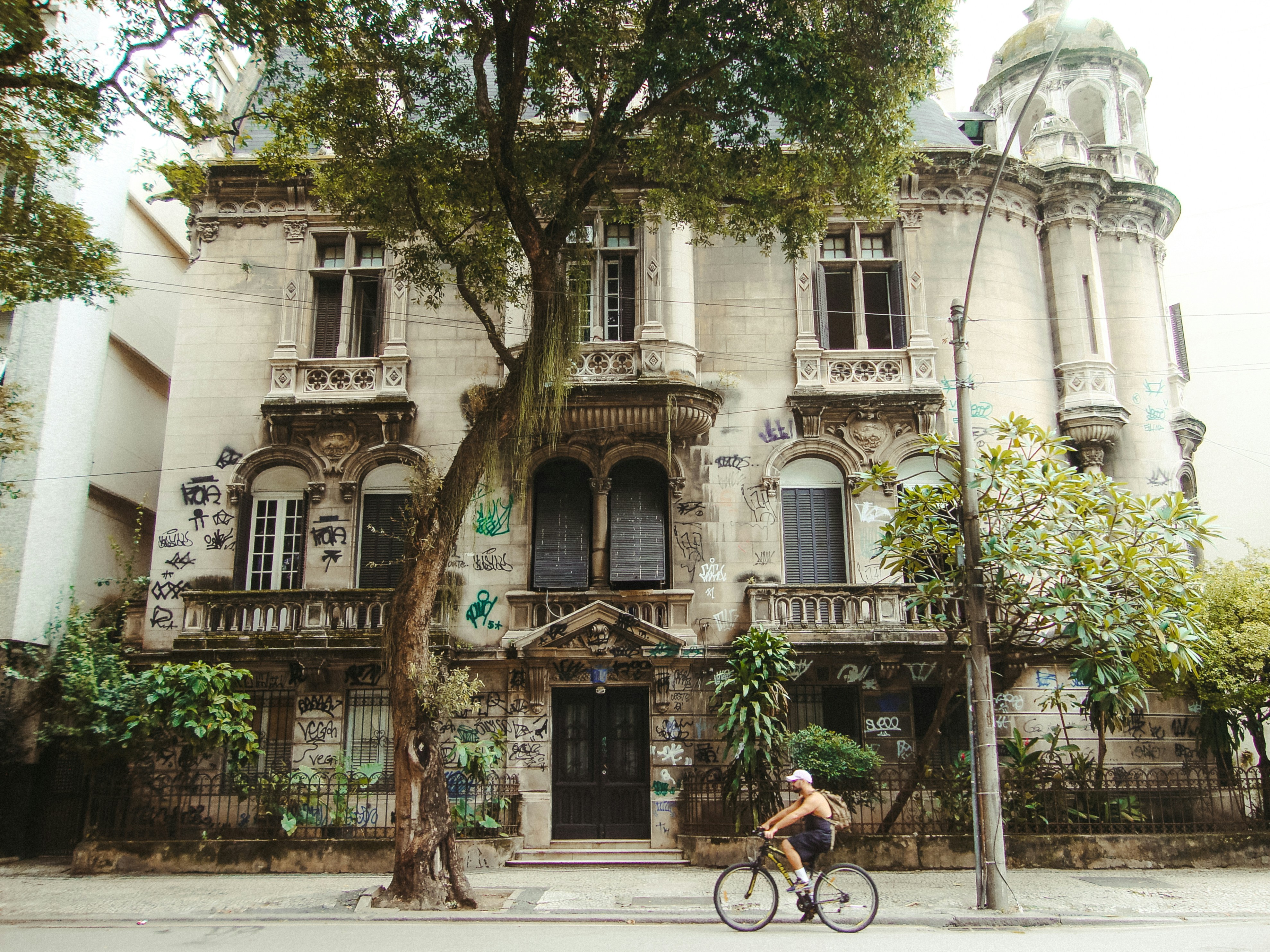Man cycles past an old ornate building with trees.