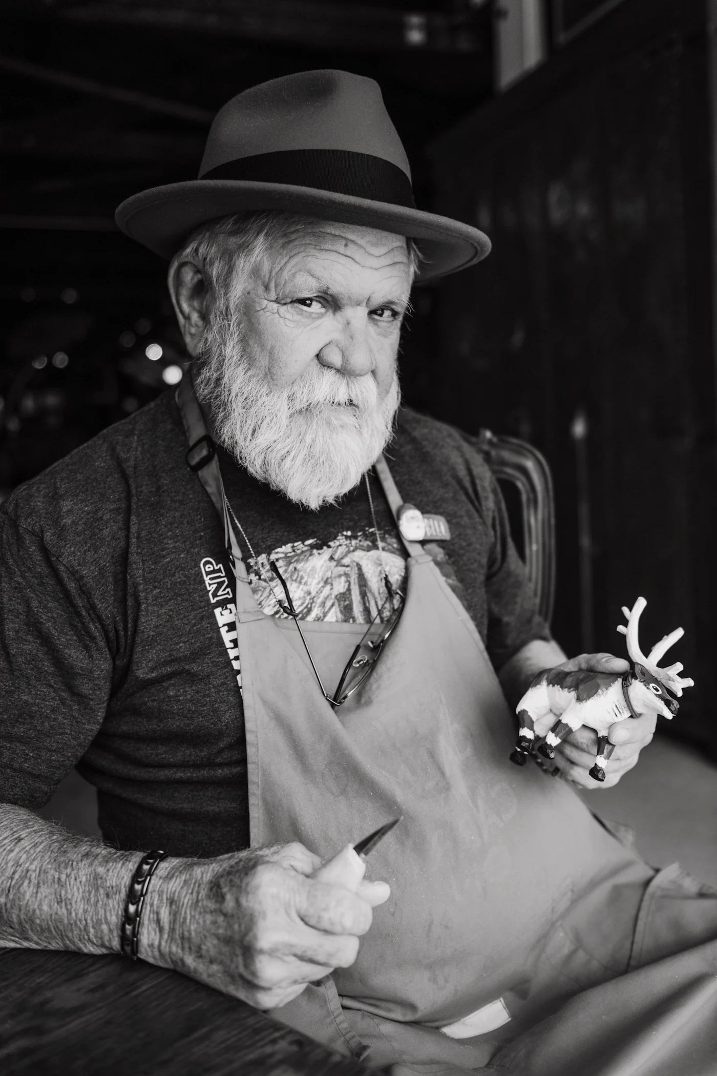 An elderly craftsman with a white beard holding carving tools and a small figure
