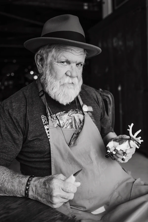 An elderly craftsman with a white beard holding carving tools and a small figure