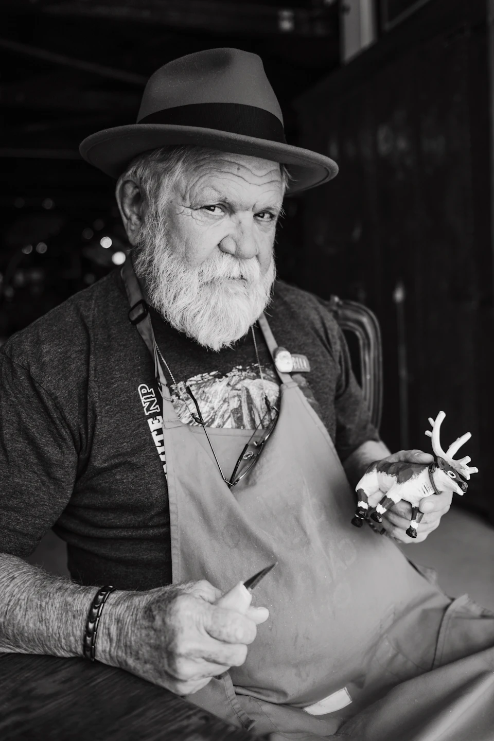 An elderly craftsman with a white beard holding carving tools and a small figure