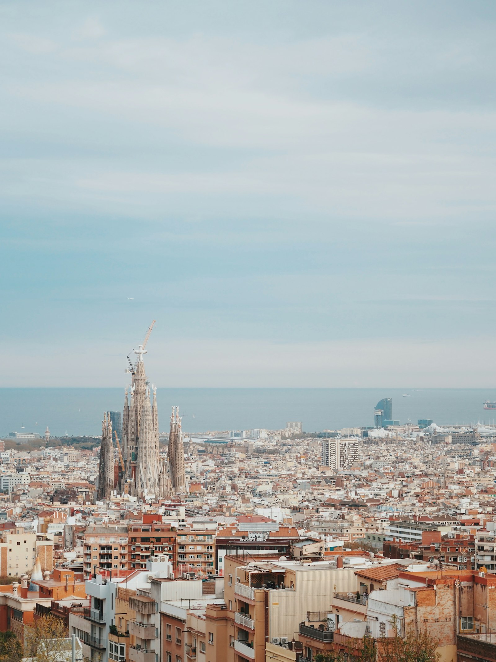 View over Barcelona's dense rooftops toward the Sagrada Família's cranes and towers, with the Mediterranean beyond