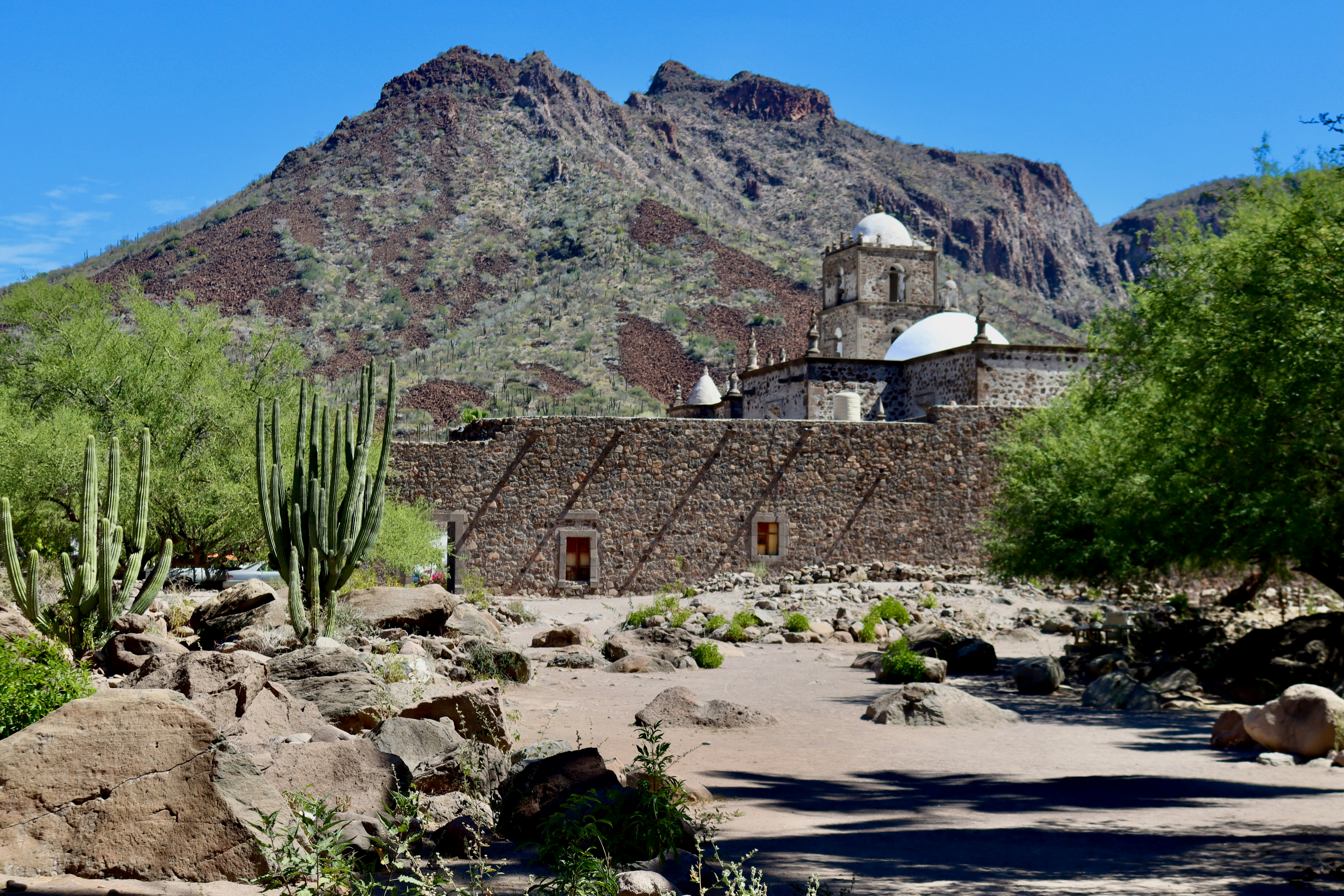 Stone building with desert plants and mountains behind.