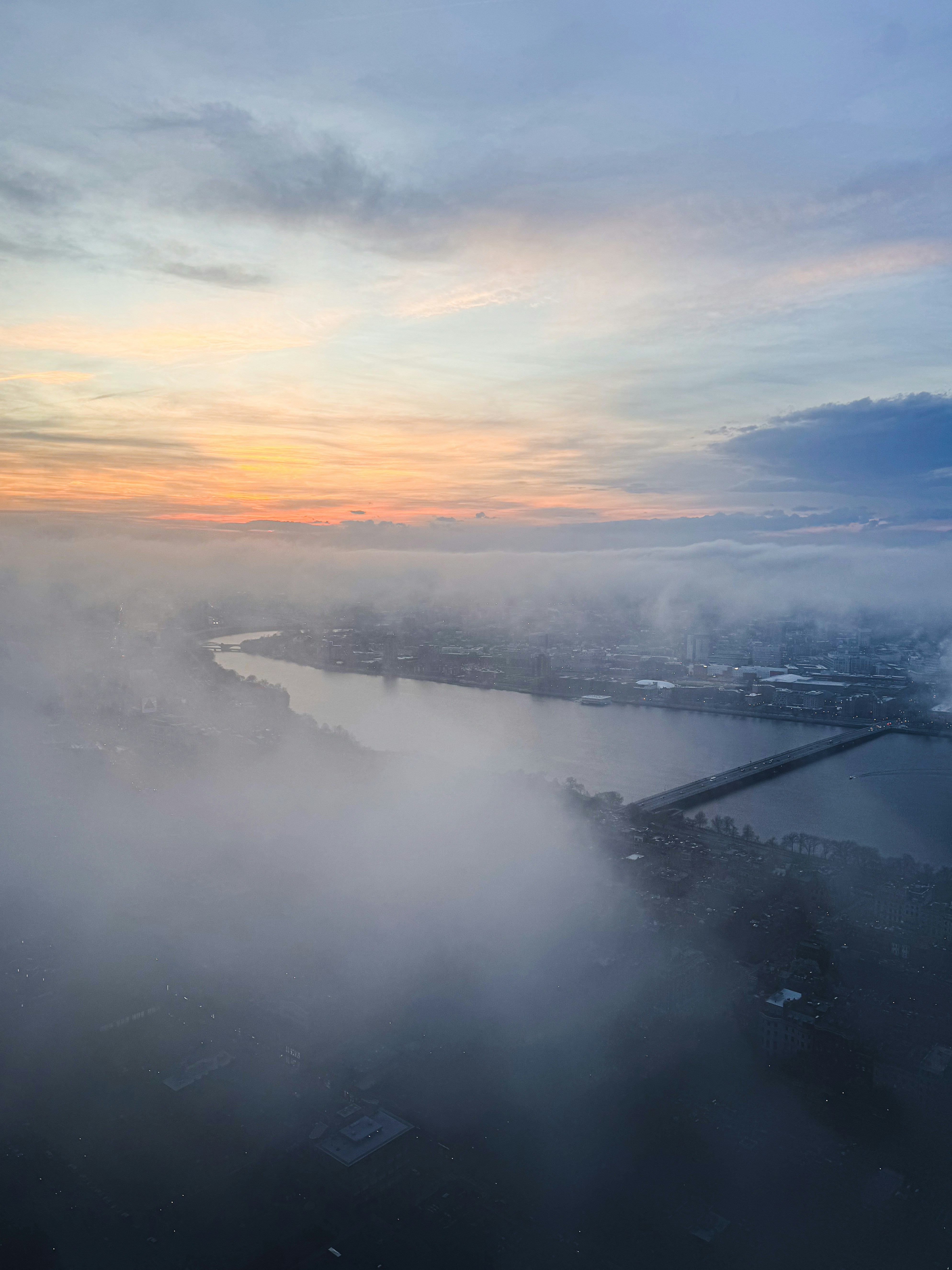 Paisaje urbano envuelto en niebla al amanecer, con río y puente.