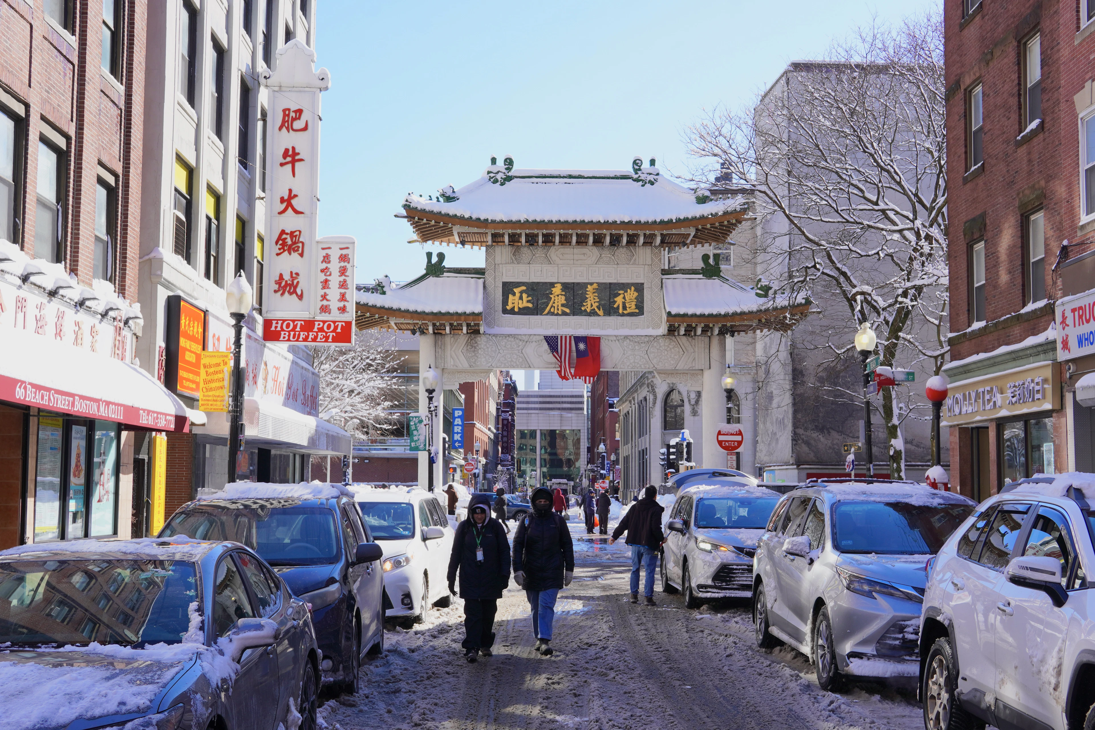 Snow-covered Beach Street in Boston's Chinatown, the Chinese paifang gate closing the end of the block and red and gold hot-pot and bakery signs stacked along the shopfronts.