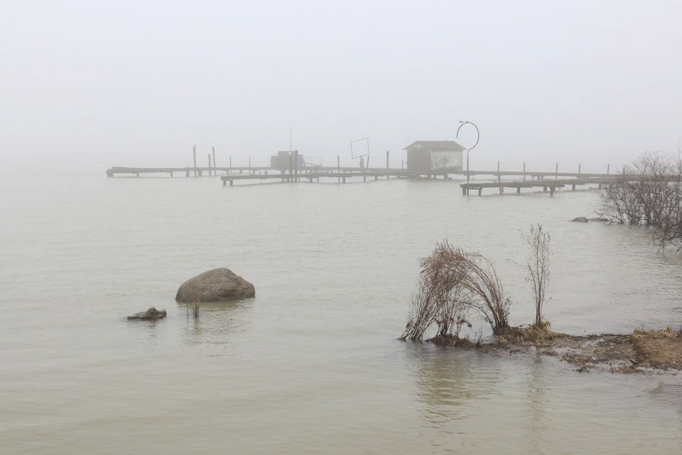 Oever van een waterplas in de mist, steigers in de verte