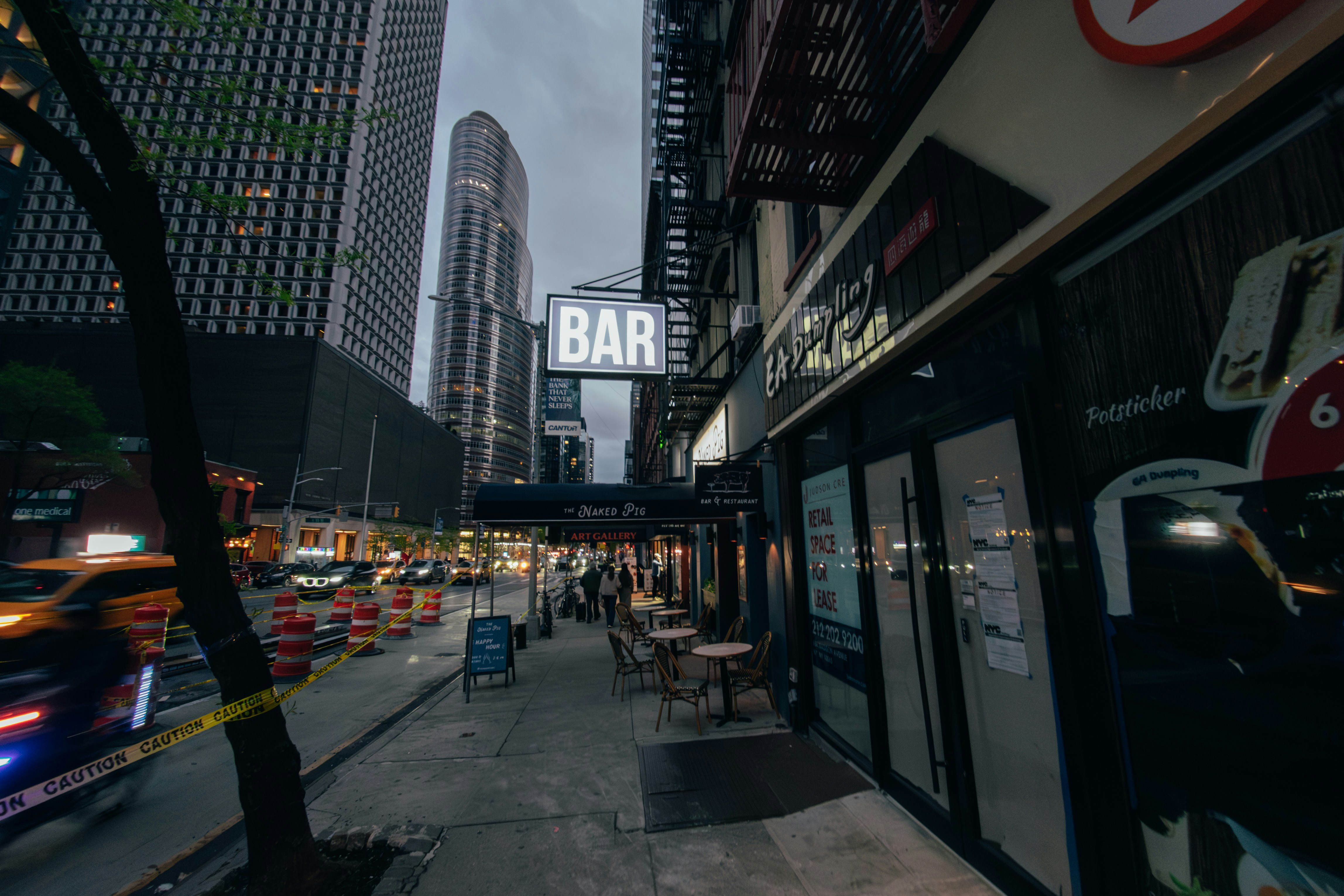 A bar sign illuminates a city street at dusk.