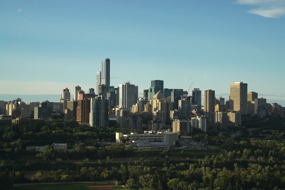 The image presents a panoramic view of the cityscape, dominated by tall buildings that stretch towards the sky. The tallest building is located on the left side of the frame and stands out with its modern design. In the center of the scene, there's a large white building with a distinctive curved roof, which appears to be a prominent landmark in the city. Surrounding this central structure are several other buildings, varying in height and architectural style. The city is nestled amidst lush green trees, providing a refreshing contrast to the urban landscape.