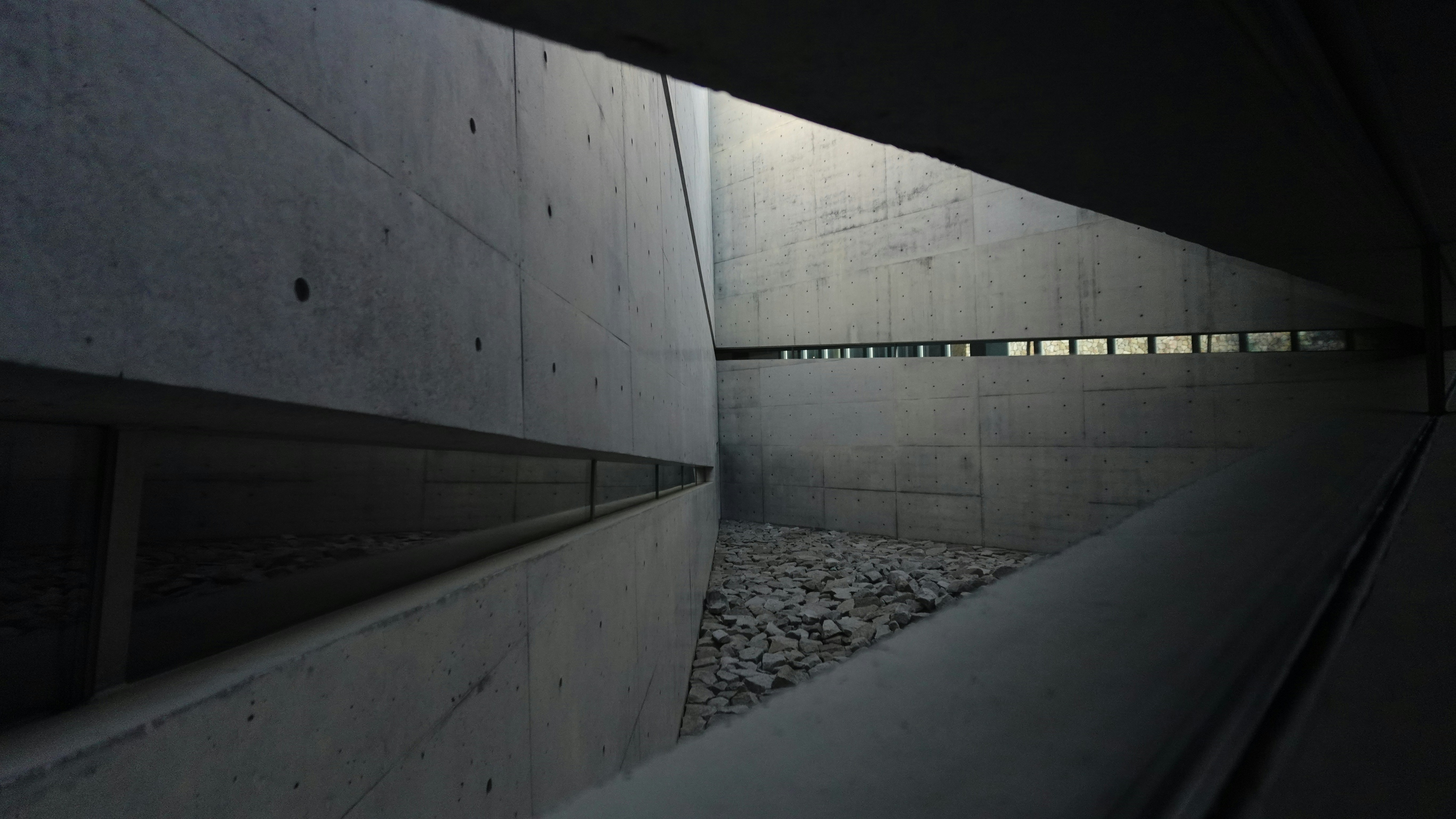 Concrete architectural corridor with stark angular planes and a pebbled ground, emphasizing lines and negative space in a muted urban environment.