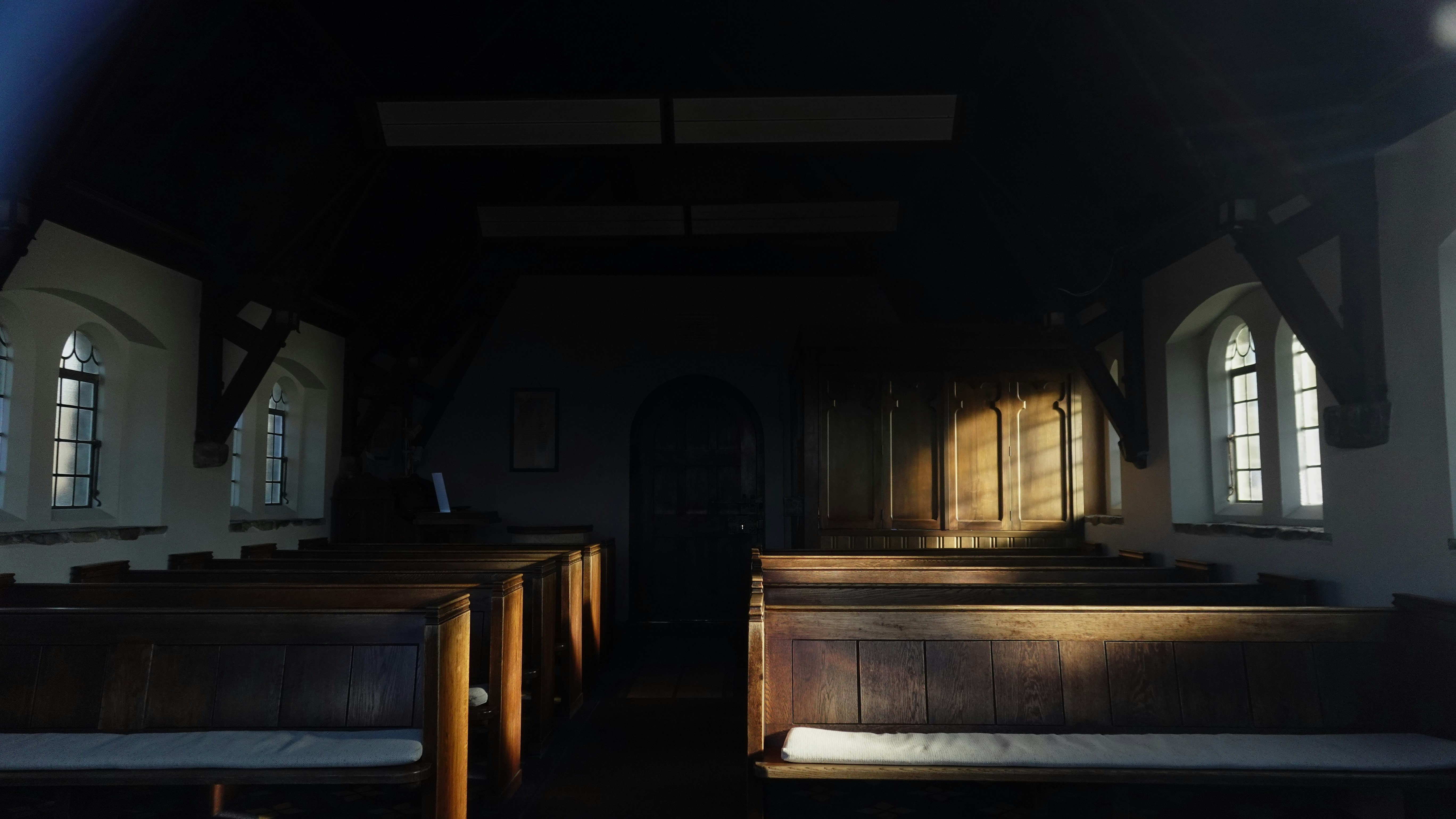 Sunlit pews line a dim church hall with warm light spilling across the wooden benches, emphasizing the central aisle and the arched doorway at the far end.