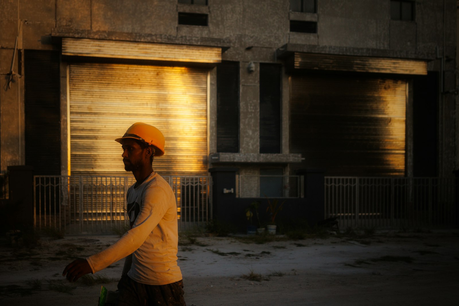 Refinery worker in a hard hat walking past process units at sunset, the working environment where flame-resistant coveralls block evaporative cooling and radiant process heat stacks on top of ambient temperature