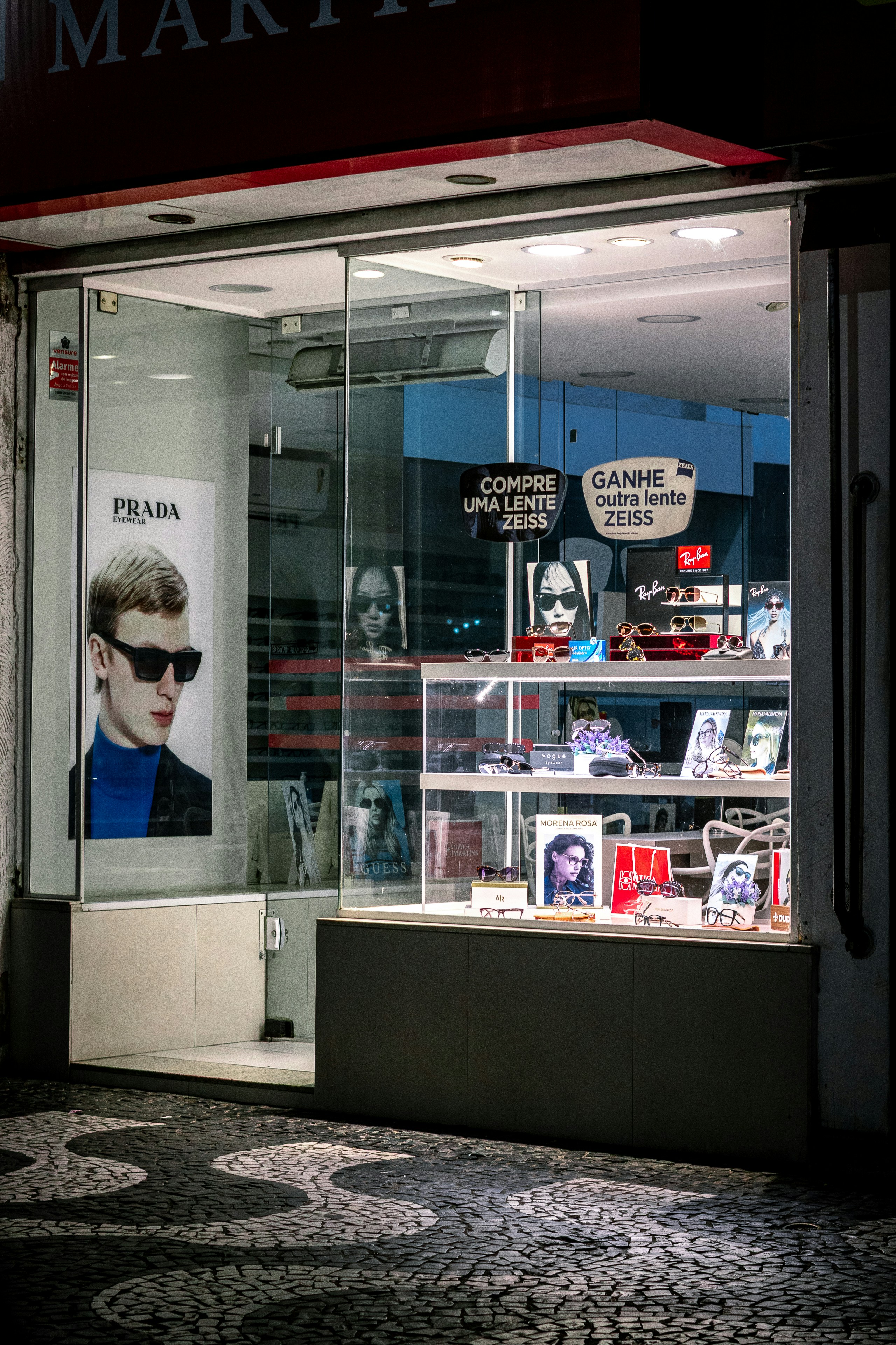 An empty, well-lit optician's shop window displays various brands of eyewear at night.