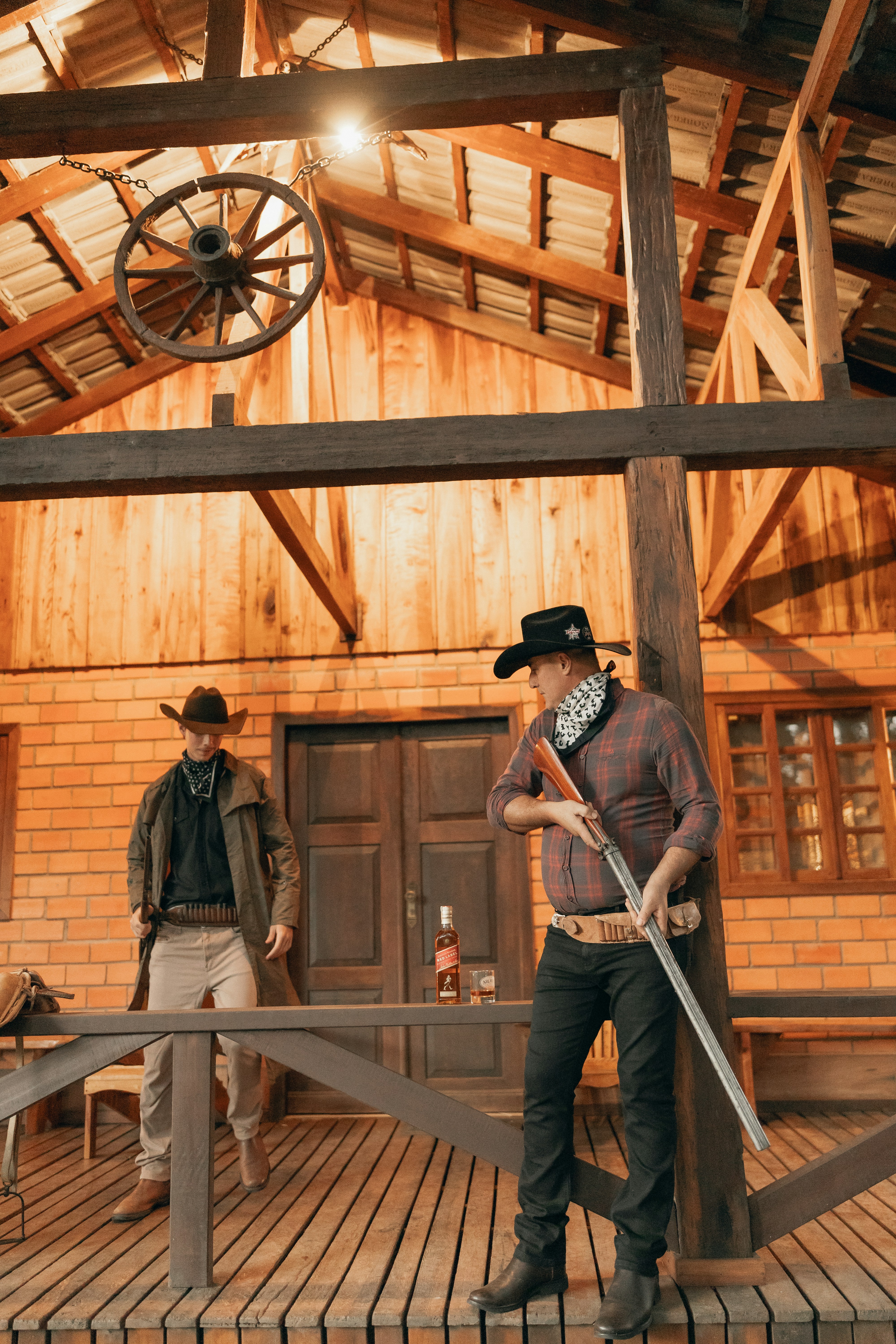 Two cowboys stand on a wooden porch with rifles