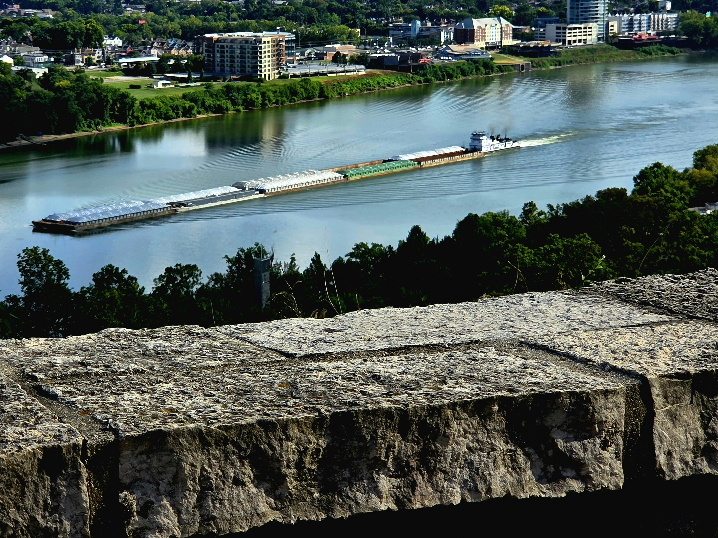 A barge travels down a wide river near a city.