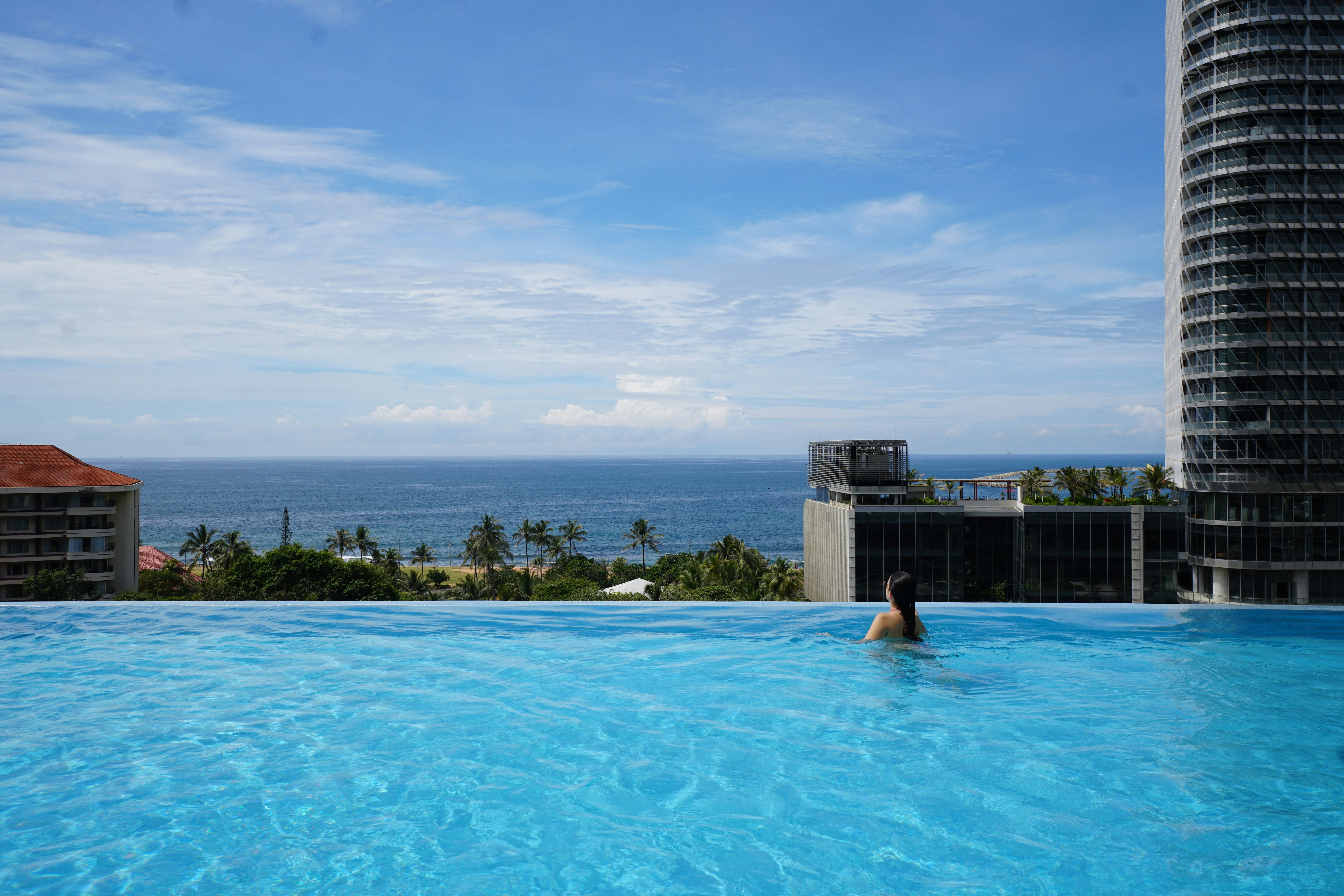 Woman swims in infinity pool overlooking ocean and cityscape