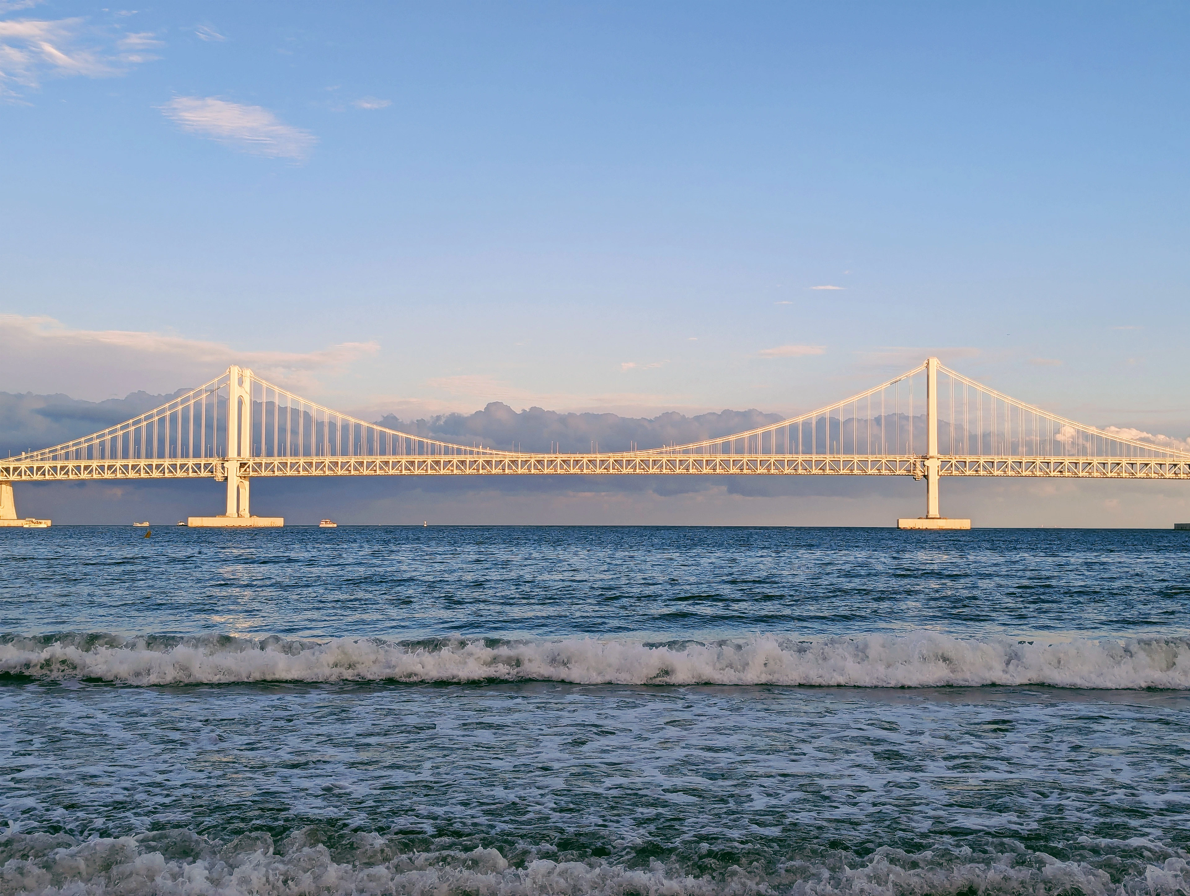 The white towers and cables of the Gwangan Bridge stretching right across the sea off Gwangalli Beach in Busan, lit warm by low evening sun, with surf breaking on the sand in the foreground.