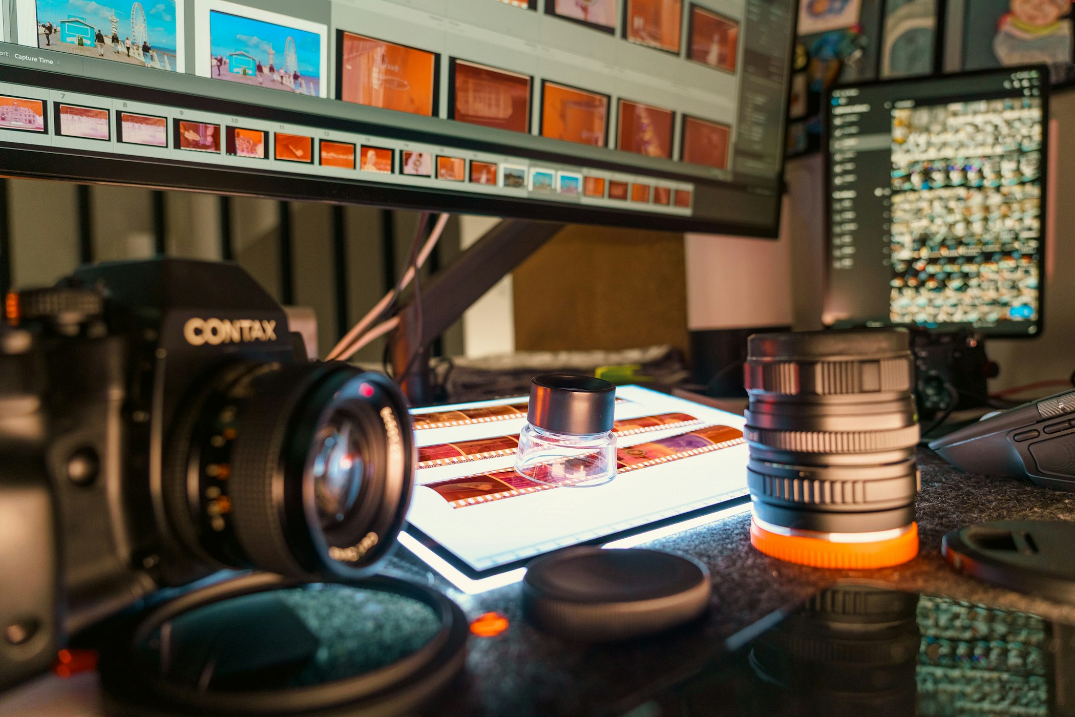 A photographer's desk with film strips on a light table, lenses, and a monitor showing a culling grid of thumbnails