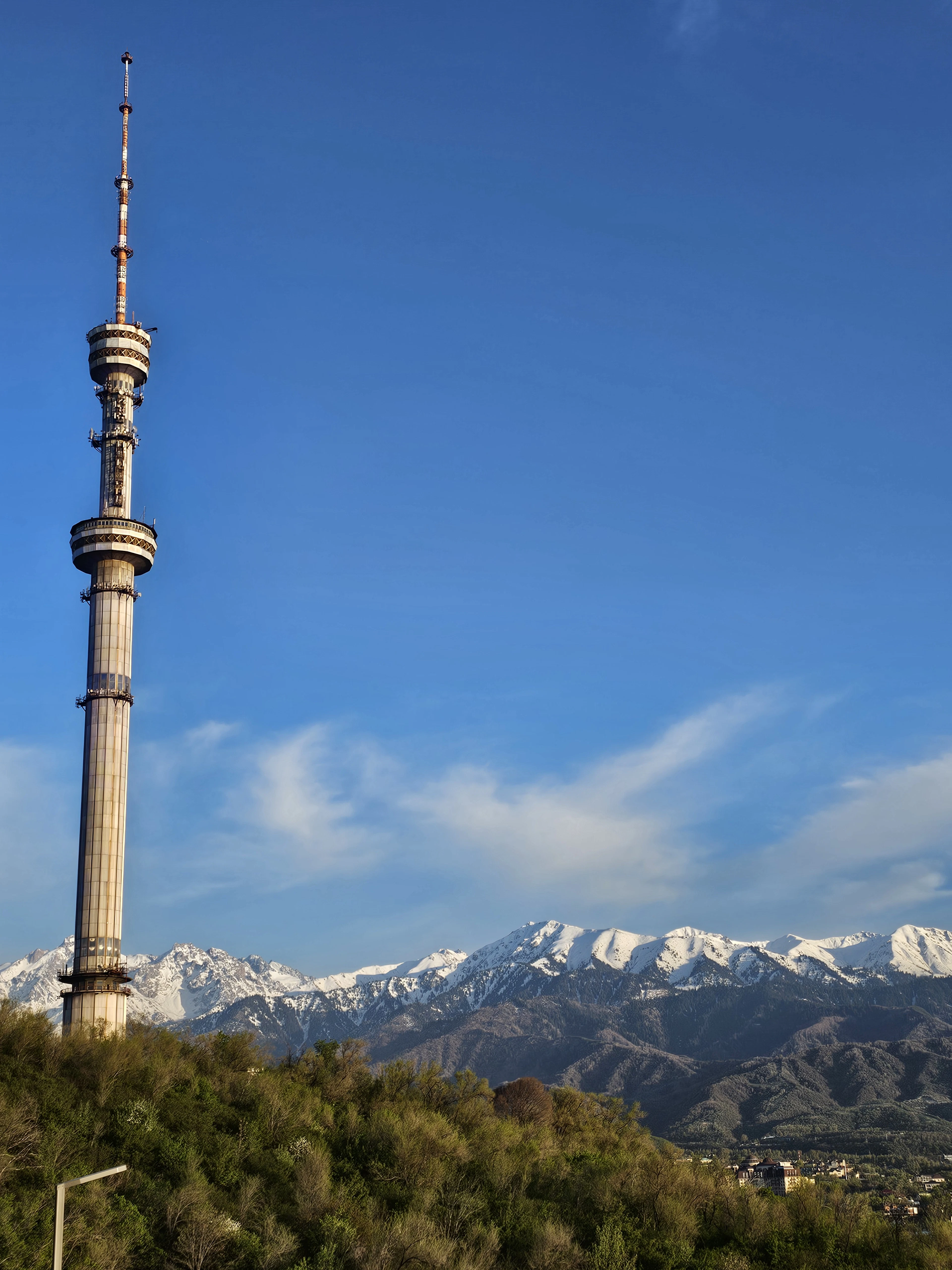 The Kok-Tobe TV tower rises above green hills, with snow-capped Tian Shan peaks and Almaty's skyline in the distance.
