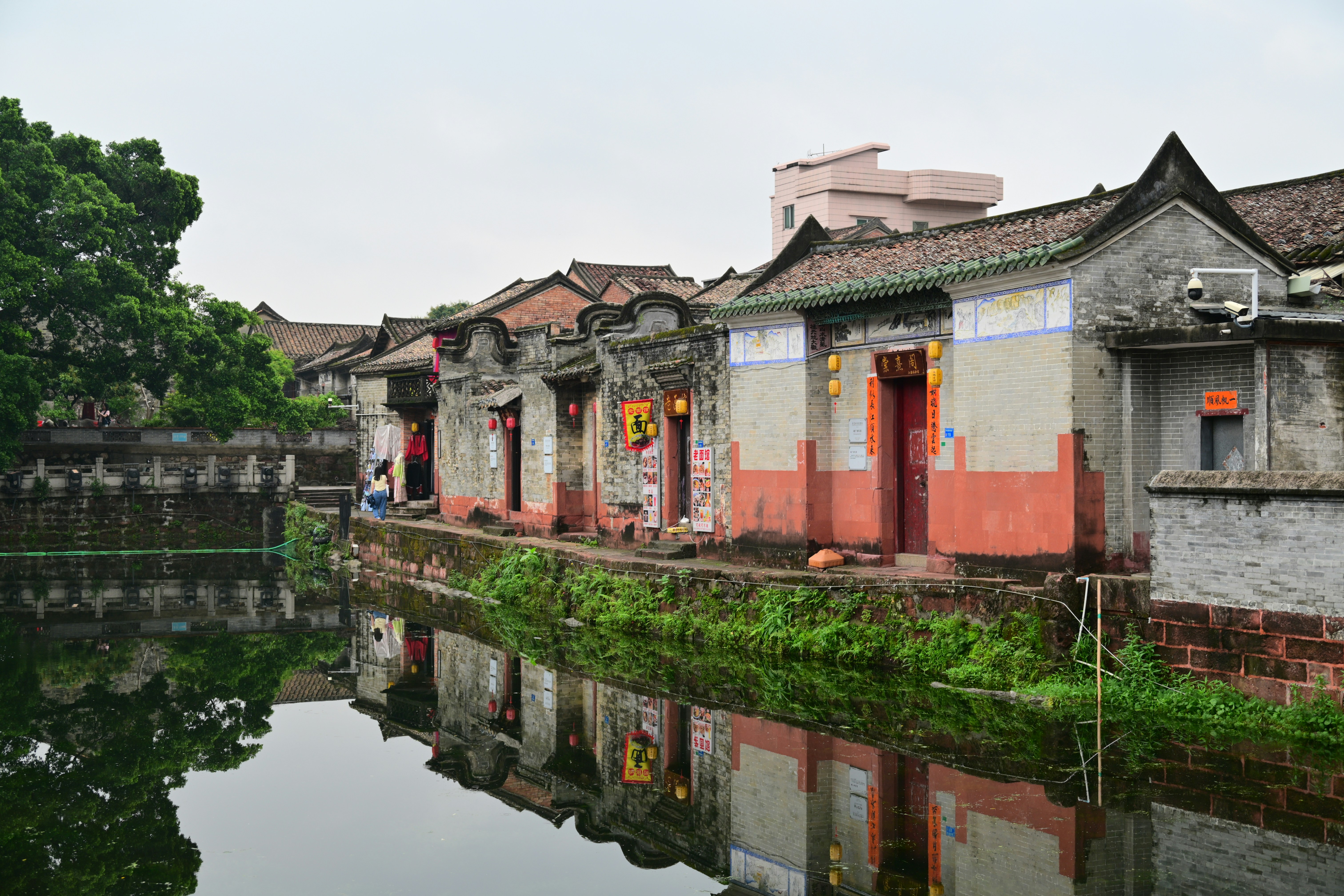 Traditional Chinese houses along a calm waterway, with their reflections mirroring in the still water under an overcast sky.