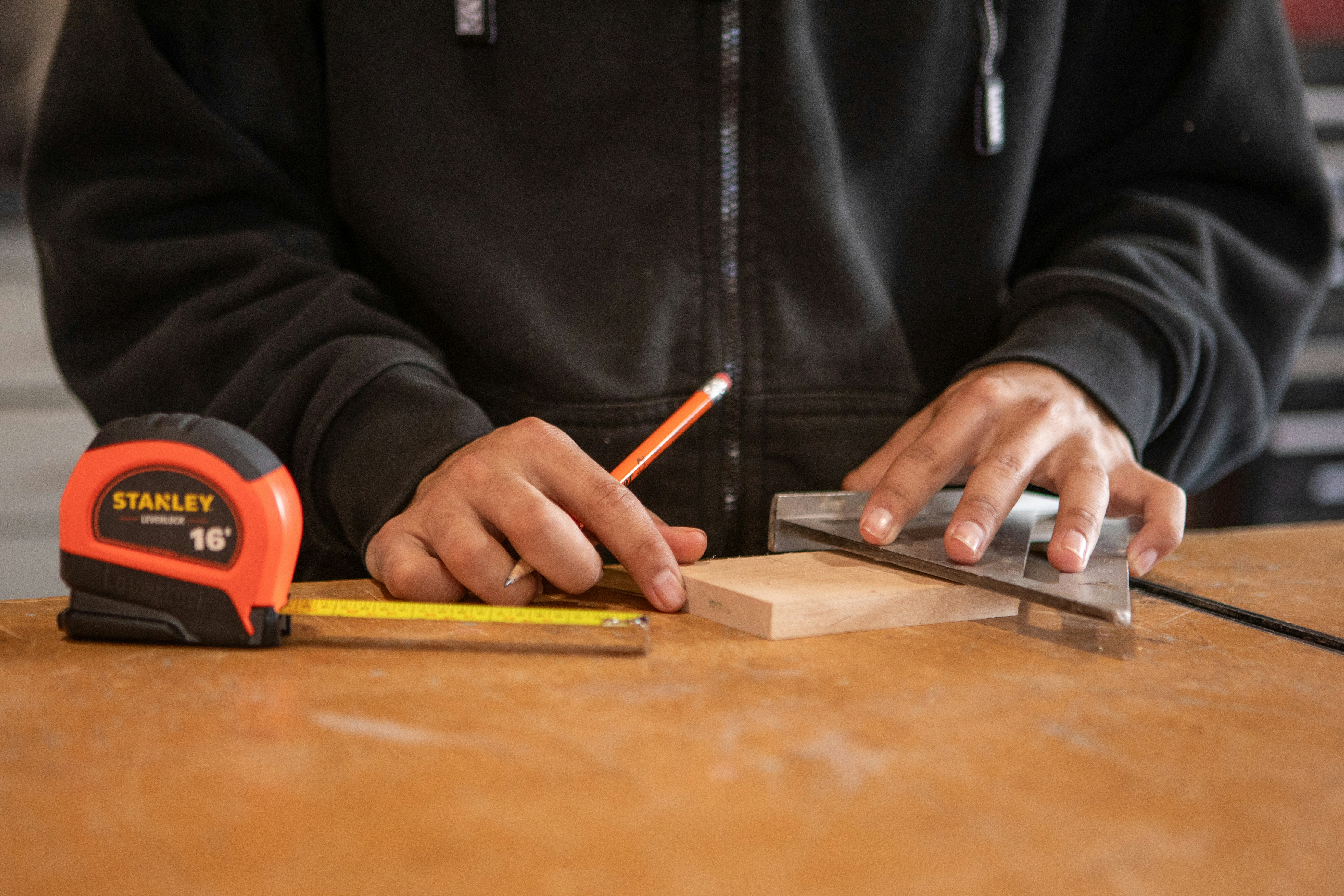Hands measuring and marking wood with pencil and square.