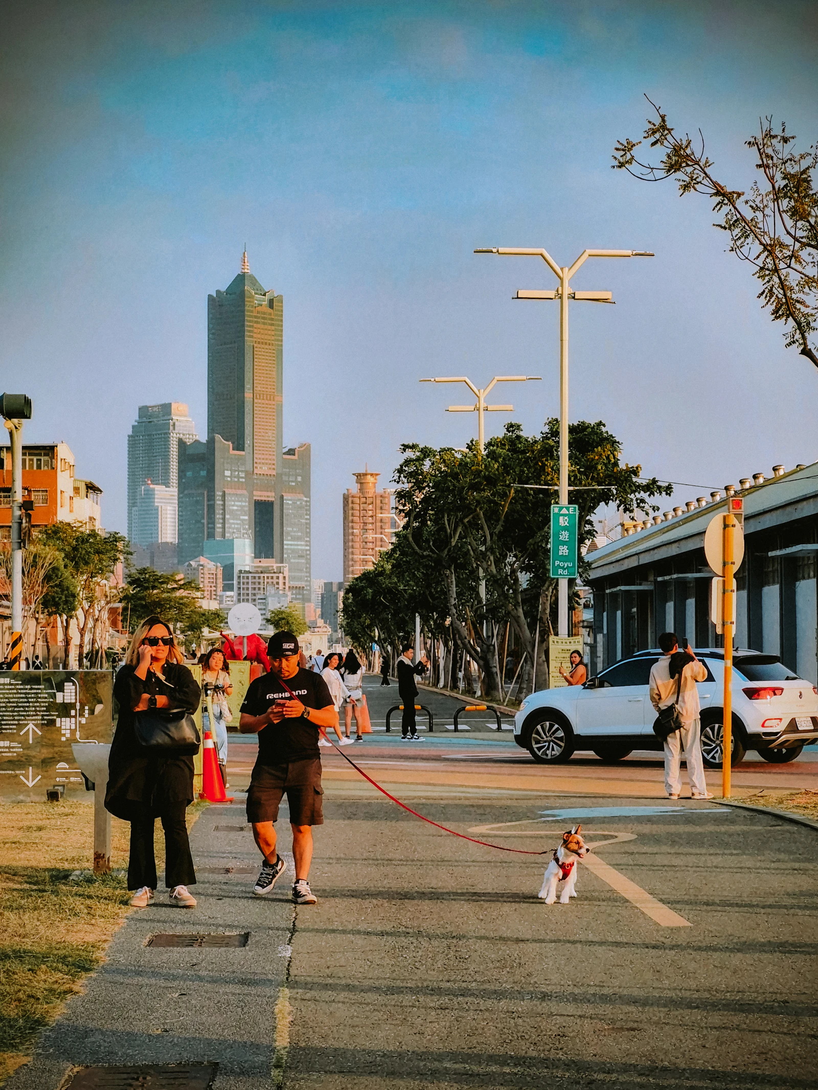 A traveler walking a small leashed dog along a waterfront path with a city skyline behind them