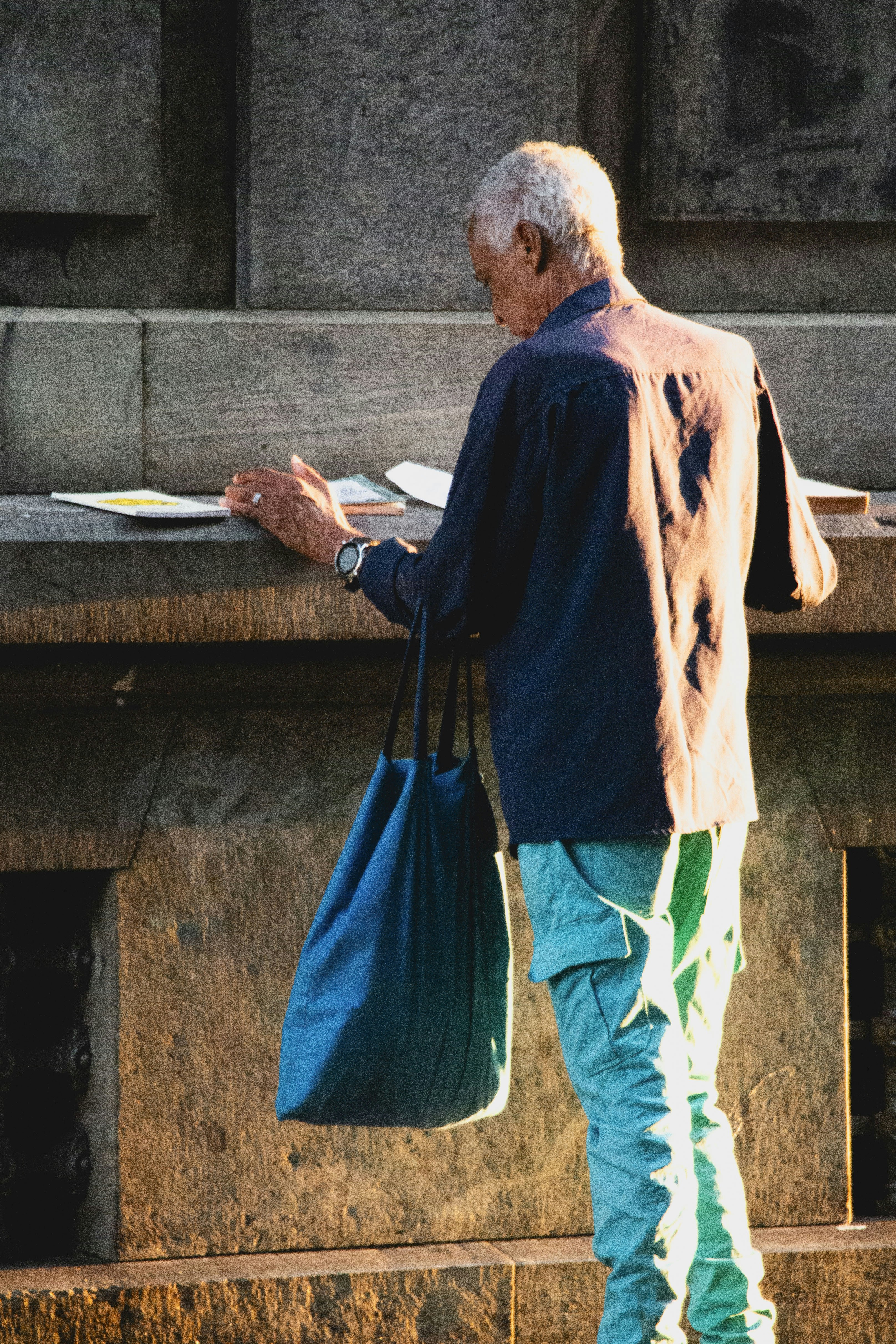 Uomo anziano con borsa blu legge vicino al muro di pietra.