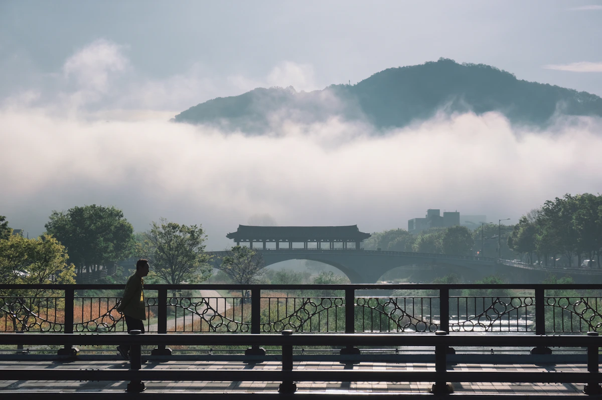 A person walks across a bridge with misty mountains behind.