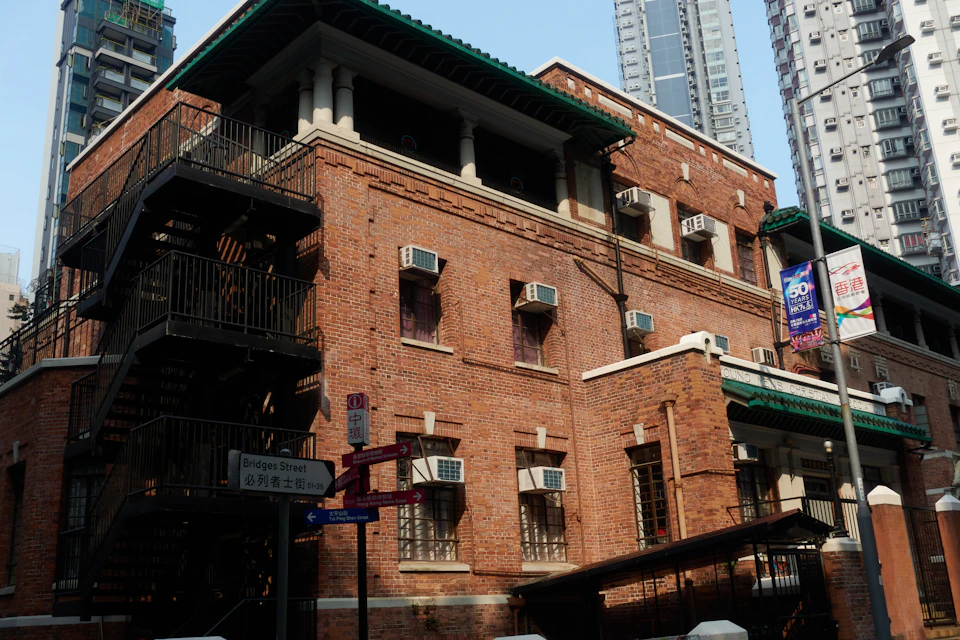 The image shows a large, two-story brick building with a green roof and multiple windows. The building has a staircase leading up to the second floor, which is accessible via an emergency exit on the ground level. A sign is visible in front of the building, though its details are not clear. In the foreground, there's a street corner with a traffic light and a pedestrian crossing sign.