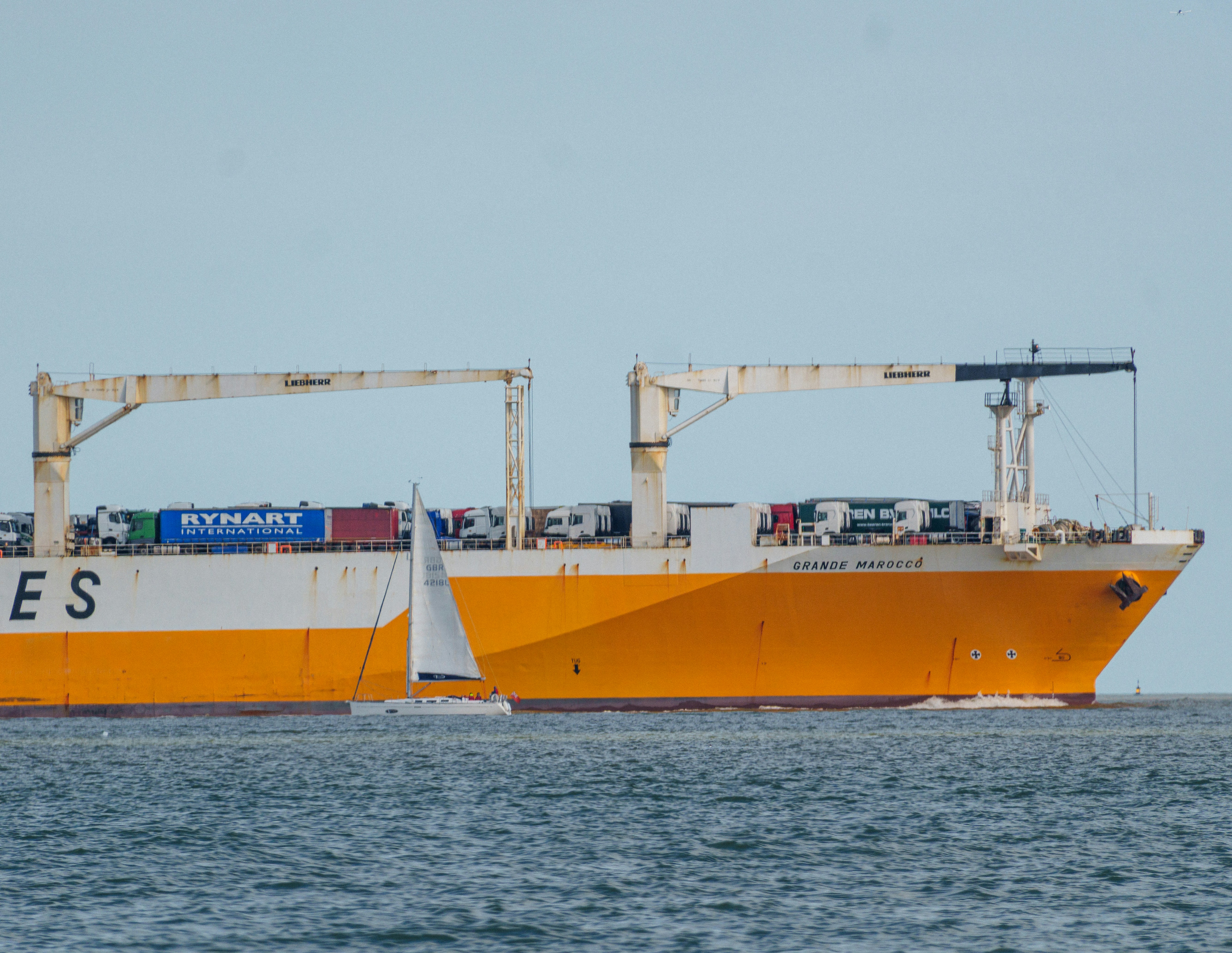 A large orange cargo ship sails past a small sailboat.
