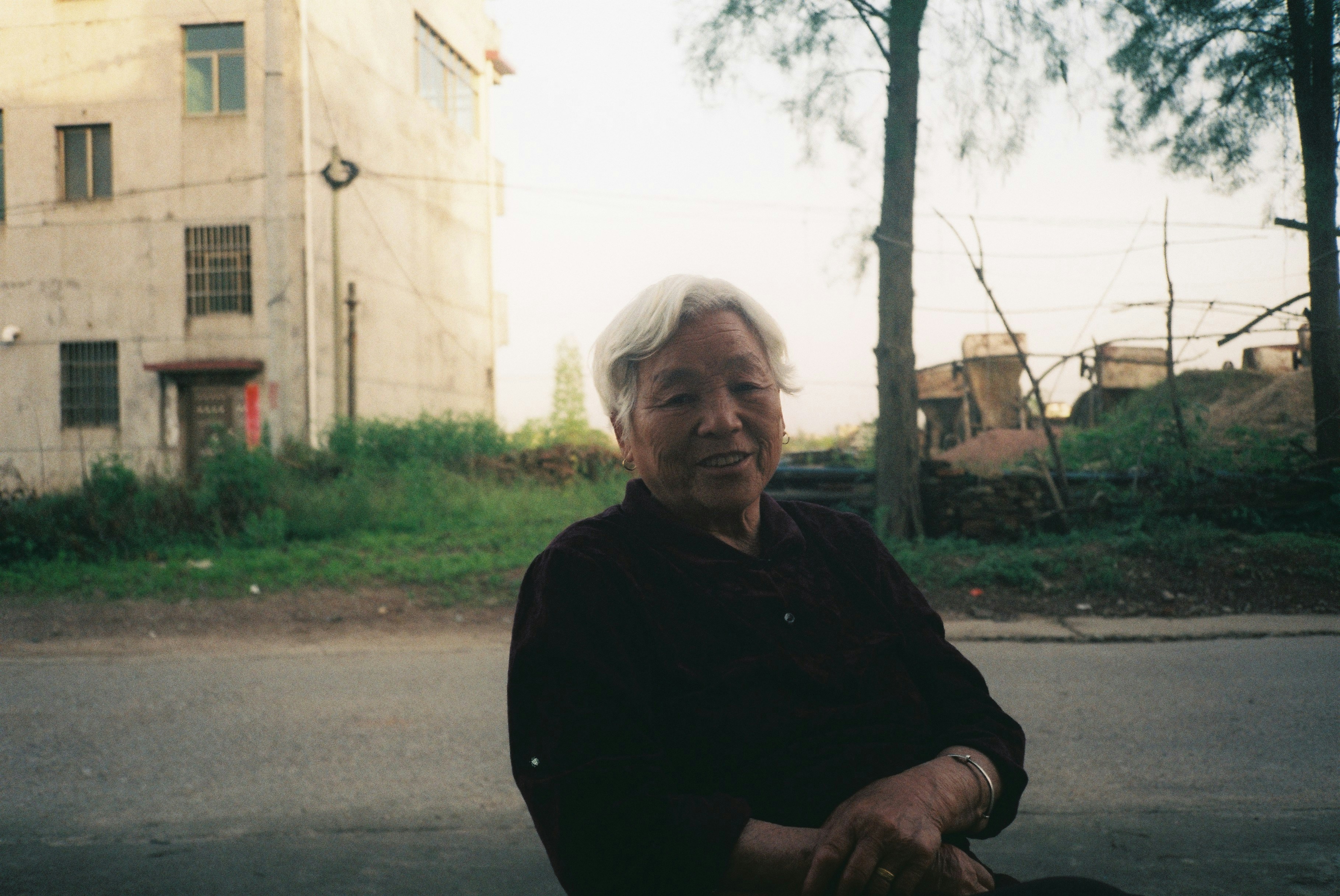 An elderly woman sits outdoors with a building behind her.