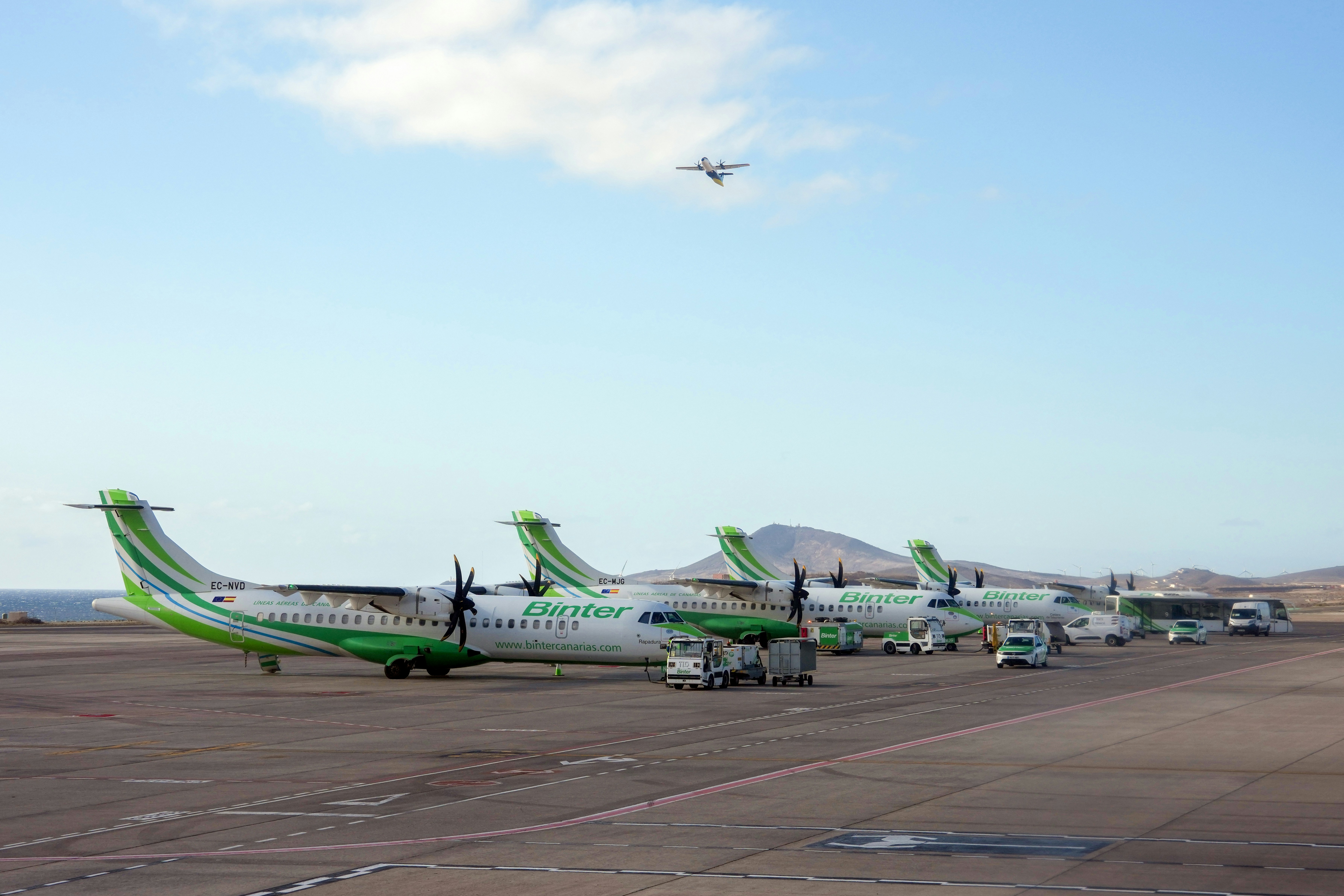 Several airplanes parked on tarmac with one flying overhead.