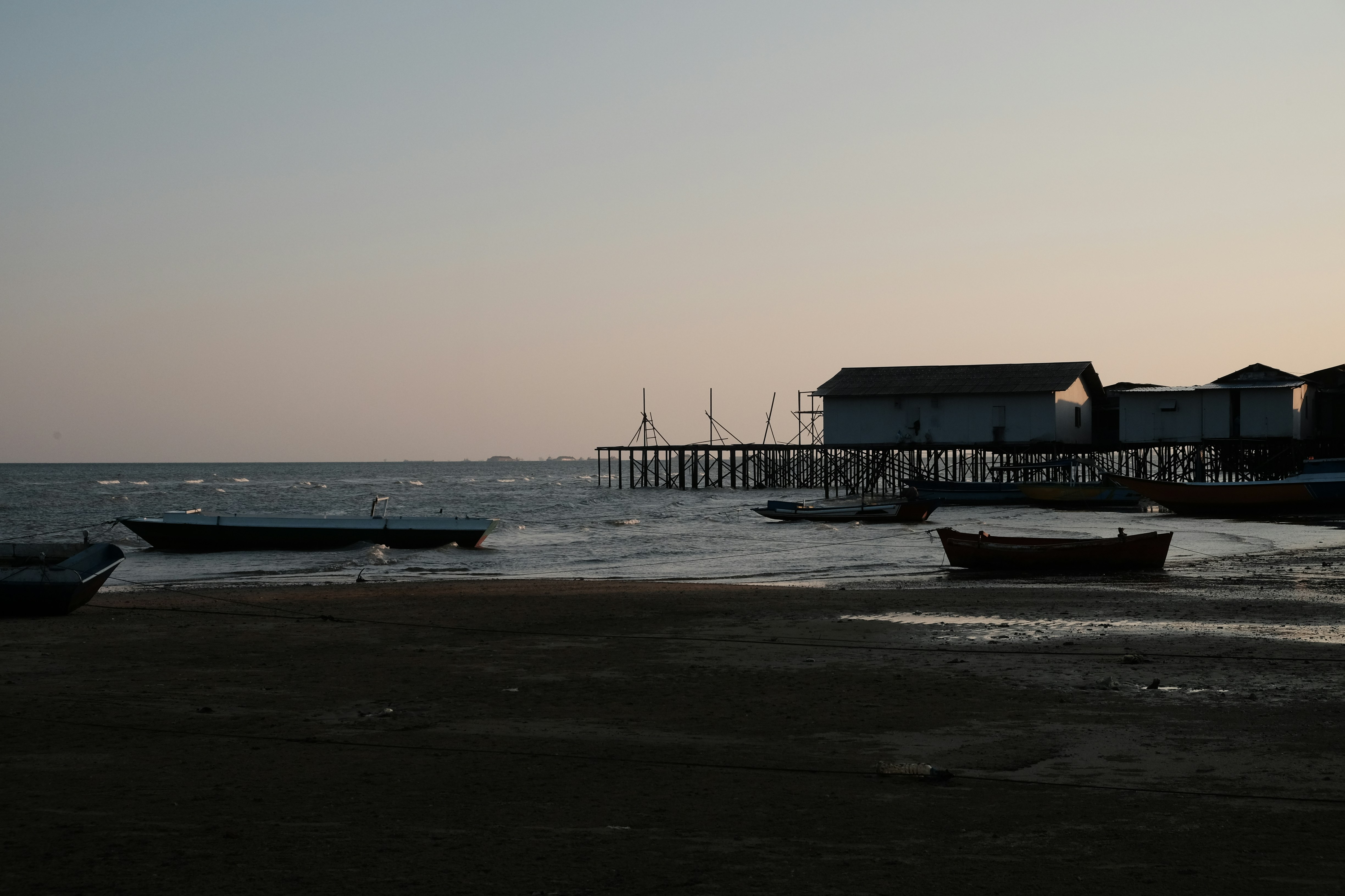 Boats rest on a muddy shore near stilted buildings.