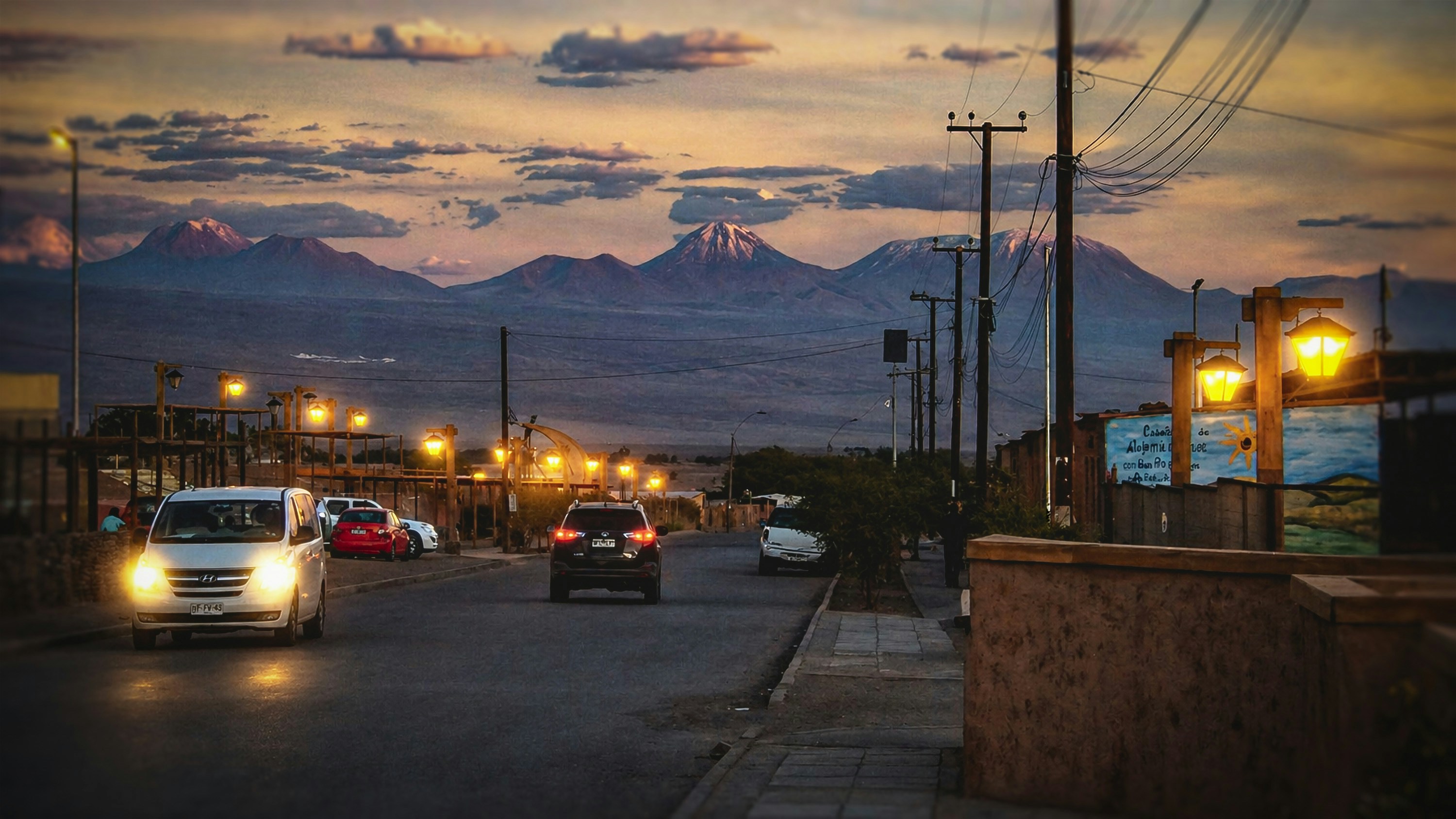 Carros passam por uma rua com montanhas ao pôr do sol.