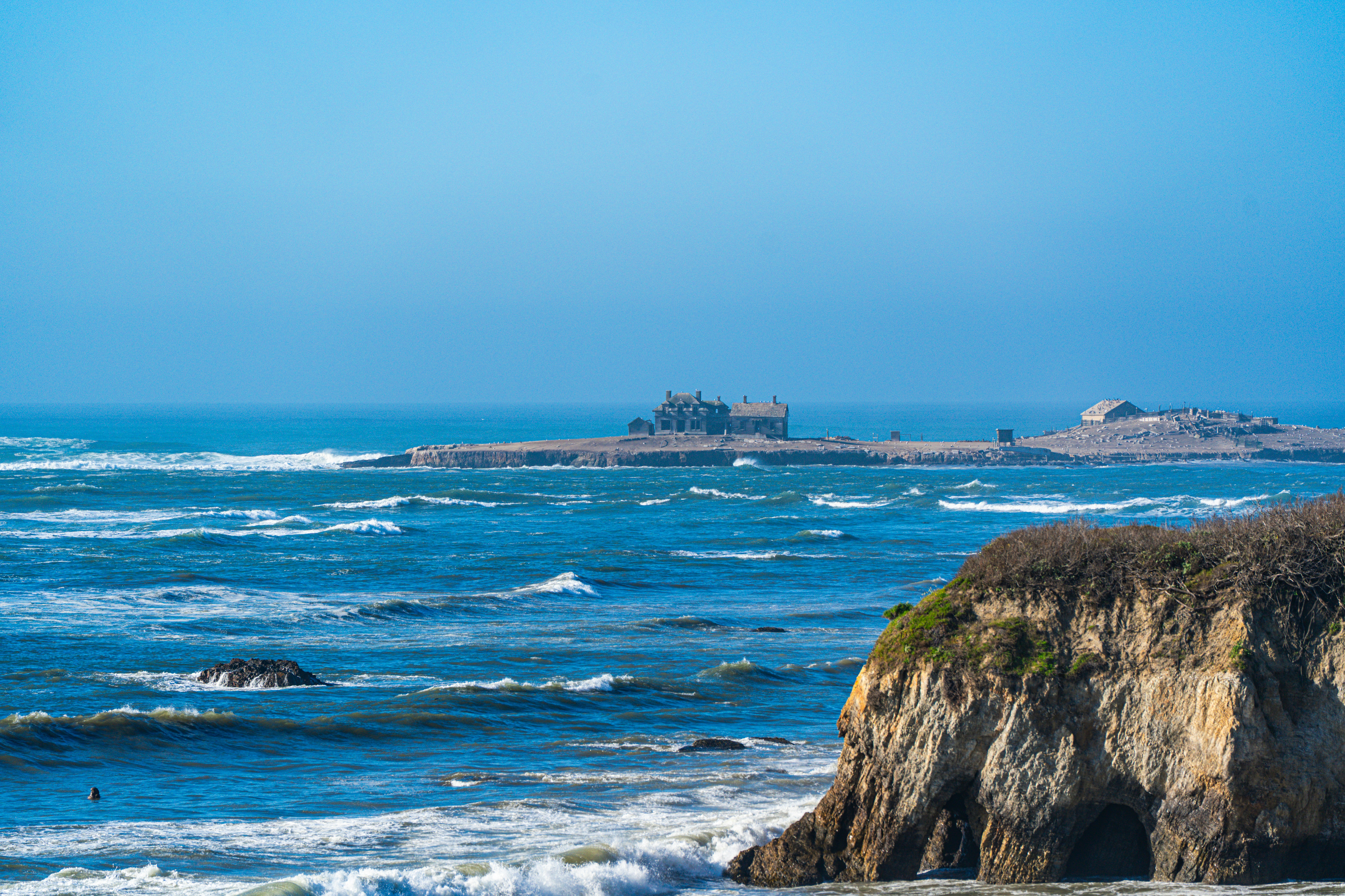 Waves crash against a rocky coastline with distant buildings.