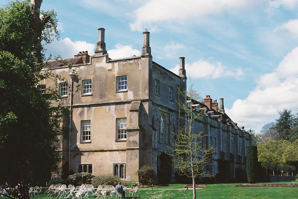 The image depicts a large, two-story stone building with multiple chimneys and a flat roof, situated on a grassy lawn surrounded by trees. The building features numerous windows that reflect the light from the clear blue sky above. In front of the building, there are several chairs arranged in a semi-circle formation, suggesting an outdoor gathering or event taking place.