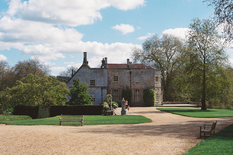 Large stone house with red roof nestled in lush greenery amidst multiple windows and chimney