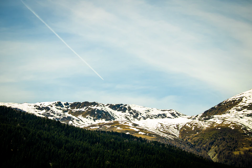 The image depicts a breathtaking mountain range under a clear blue sky with wispy clouds. The mountains, blanketed in snow, rise majestically against the horizon, their peaks touching the sky. The perspective is from below, looking up at the towering heights of these natural wonders. A small trail winds its way through the mountains, disappearing into the distance. The image does not contain any discernible text or countable objects, and there are no people or animals visible in this serene landscape.