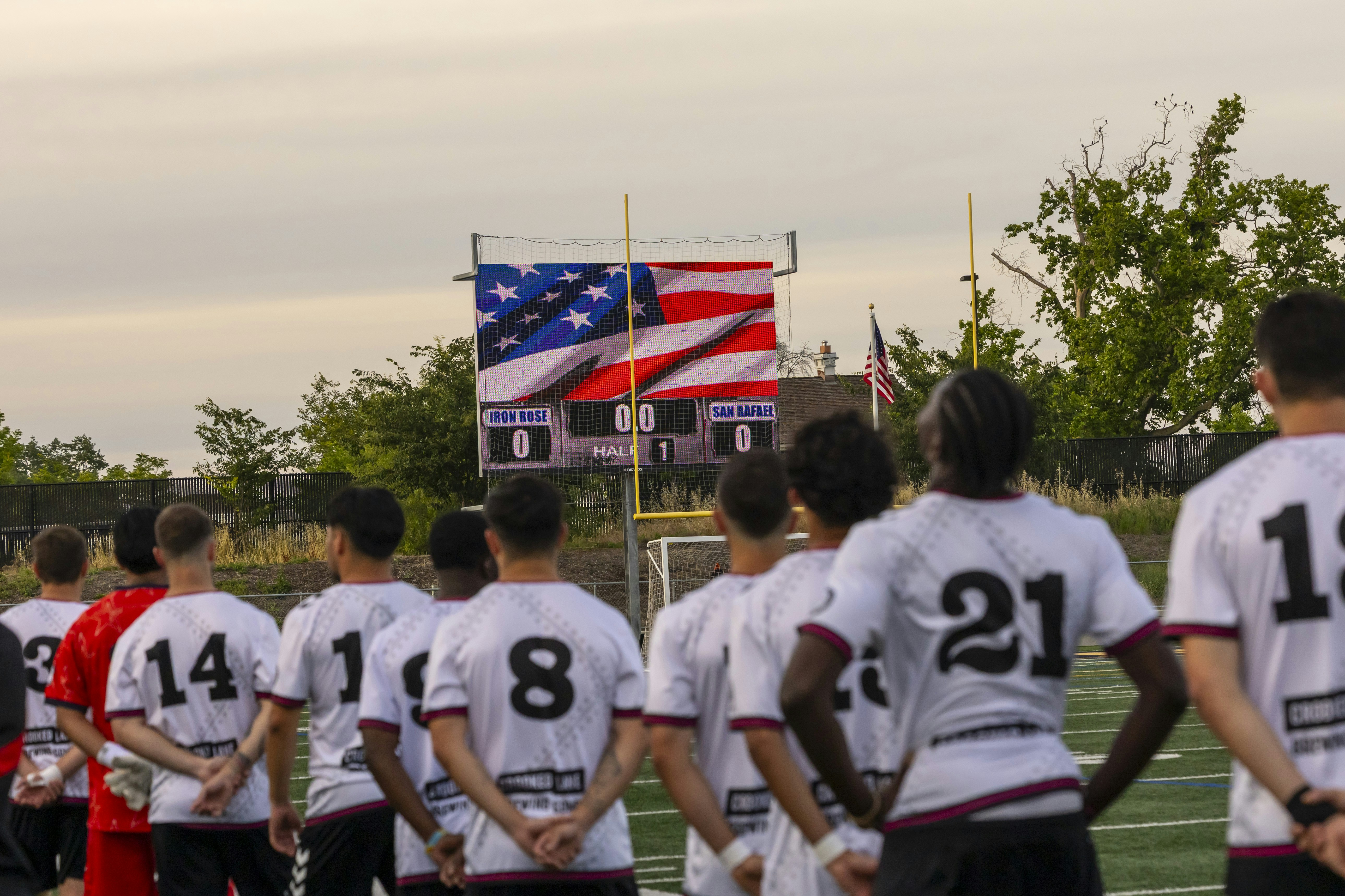 Soccer players stand facing a scoreboard with american flag.