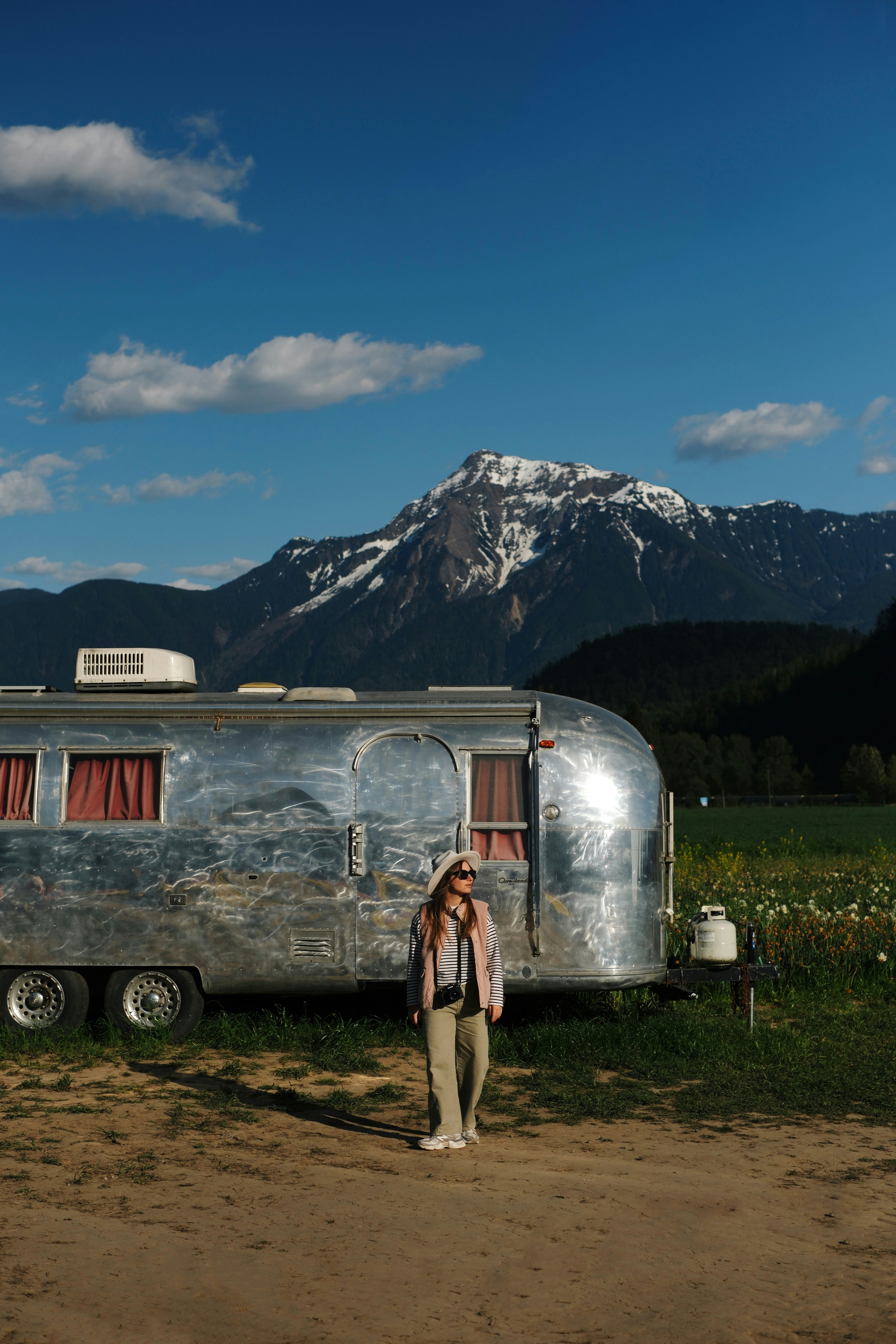 Woman standing by a vintage trailer with mountains behind.