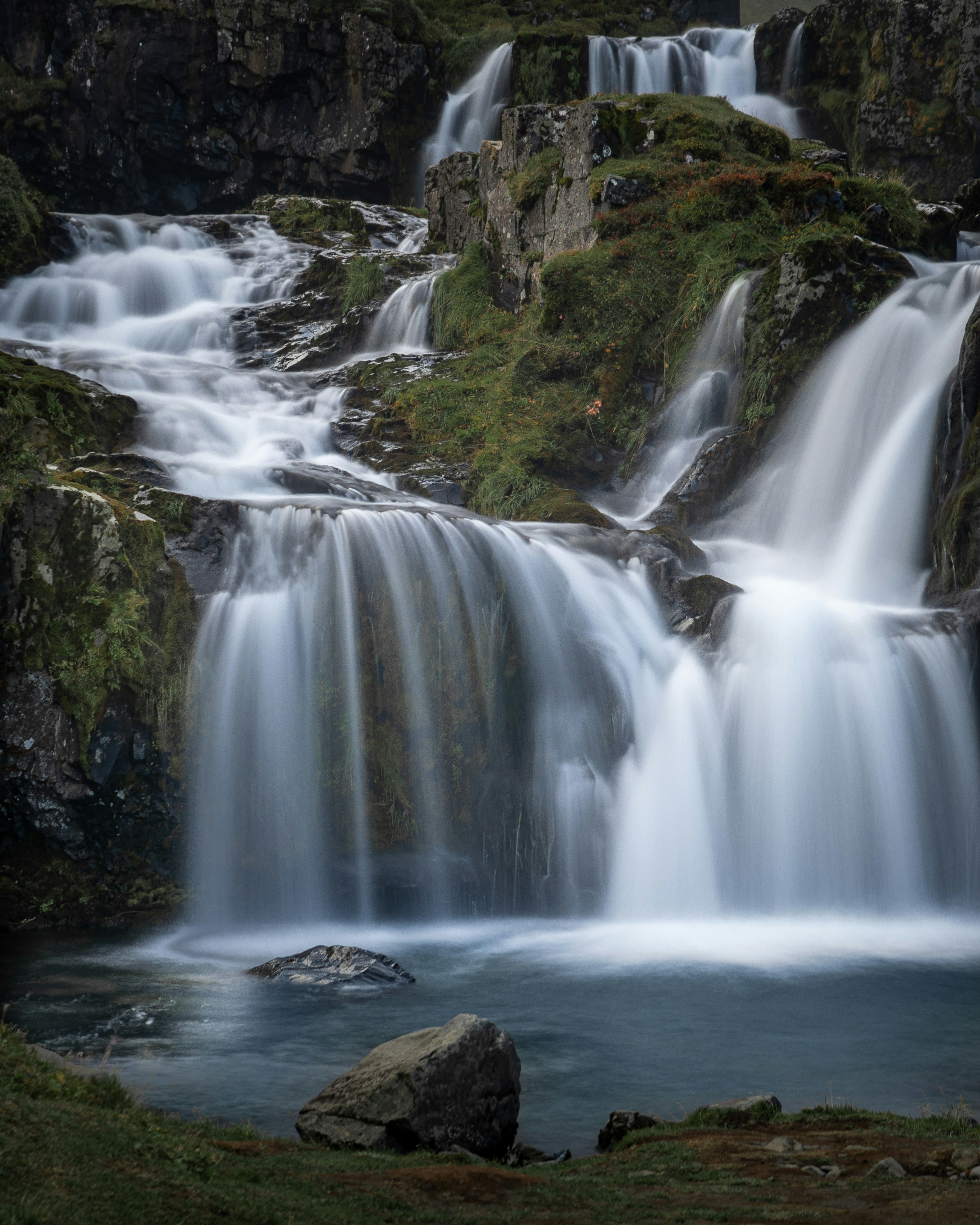 Cascading waterfalls flow over mossy rocks in a lush landscape.
