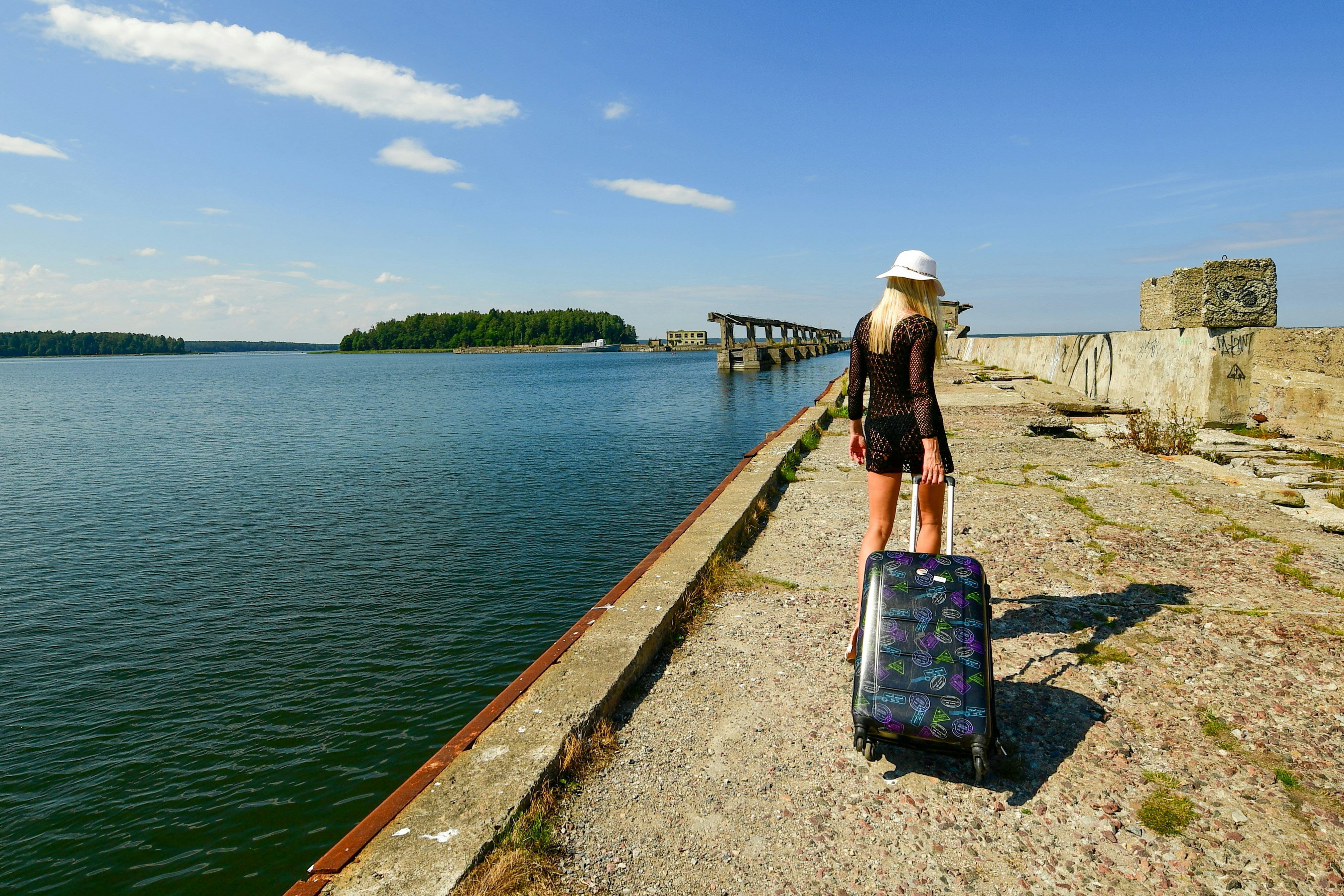 A blonde woman in a black dress and white hat walks along a concrete pier in Hara Submarine Base, pulling a large dark wheeled suitcase against a backdrop of blue sea and sky on a sunny summer day.