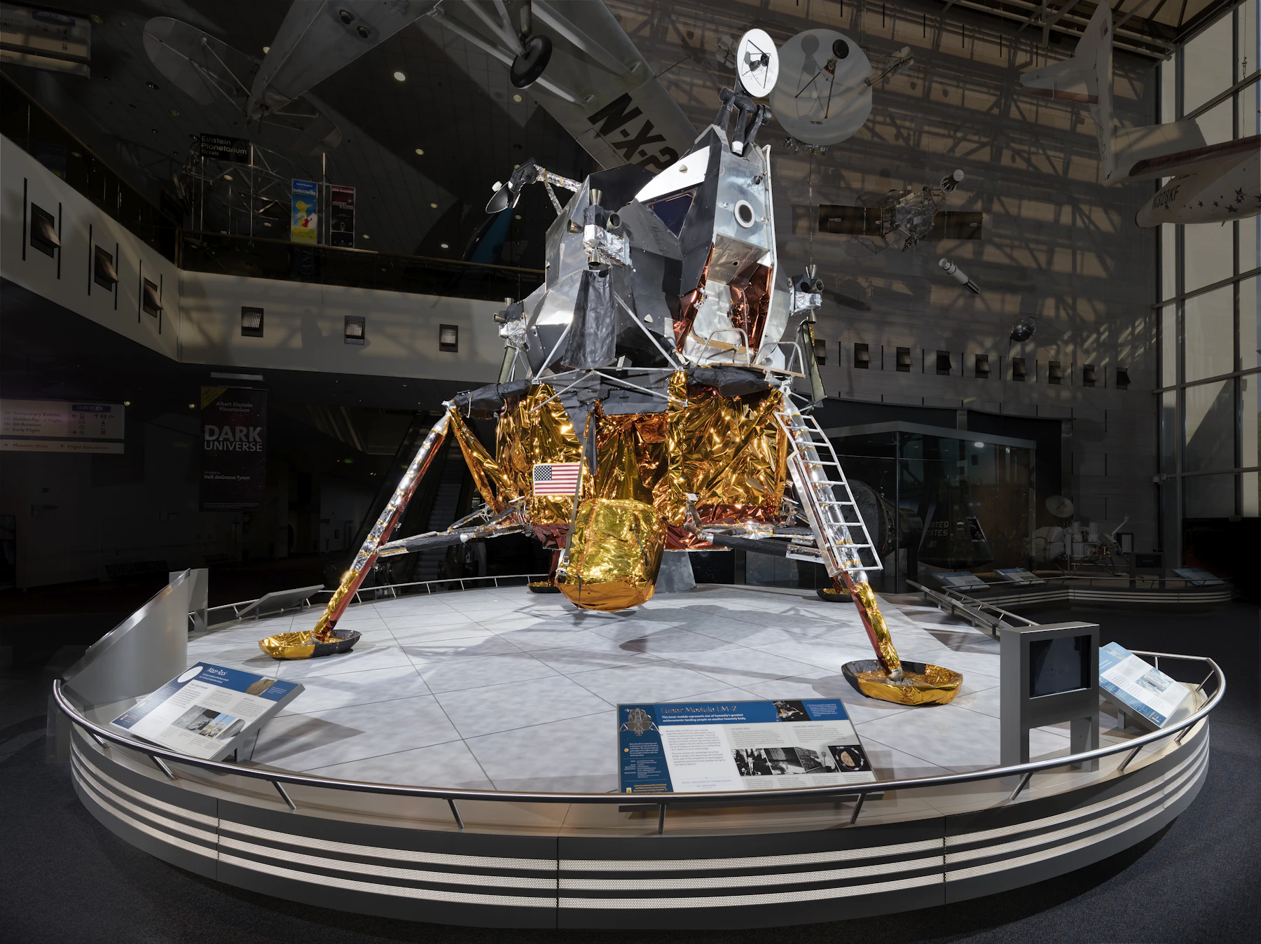 A lunar module wrapped in gold foil standing on a round platform in a museum hall
