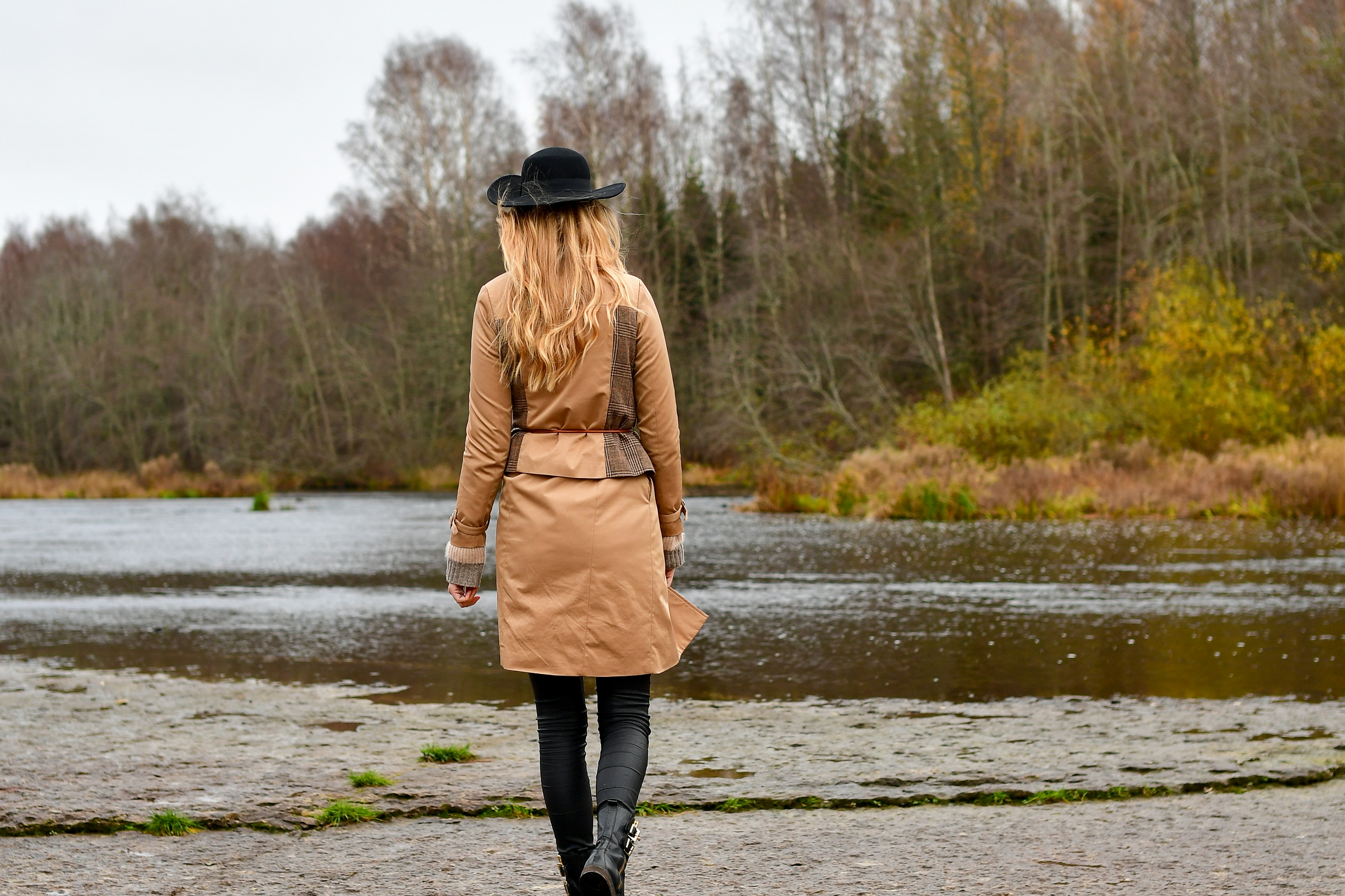Back view of a stylish woman in a trench coat and black hat walking towards a serene river in autumn.