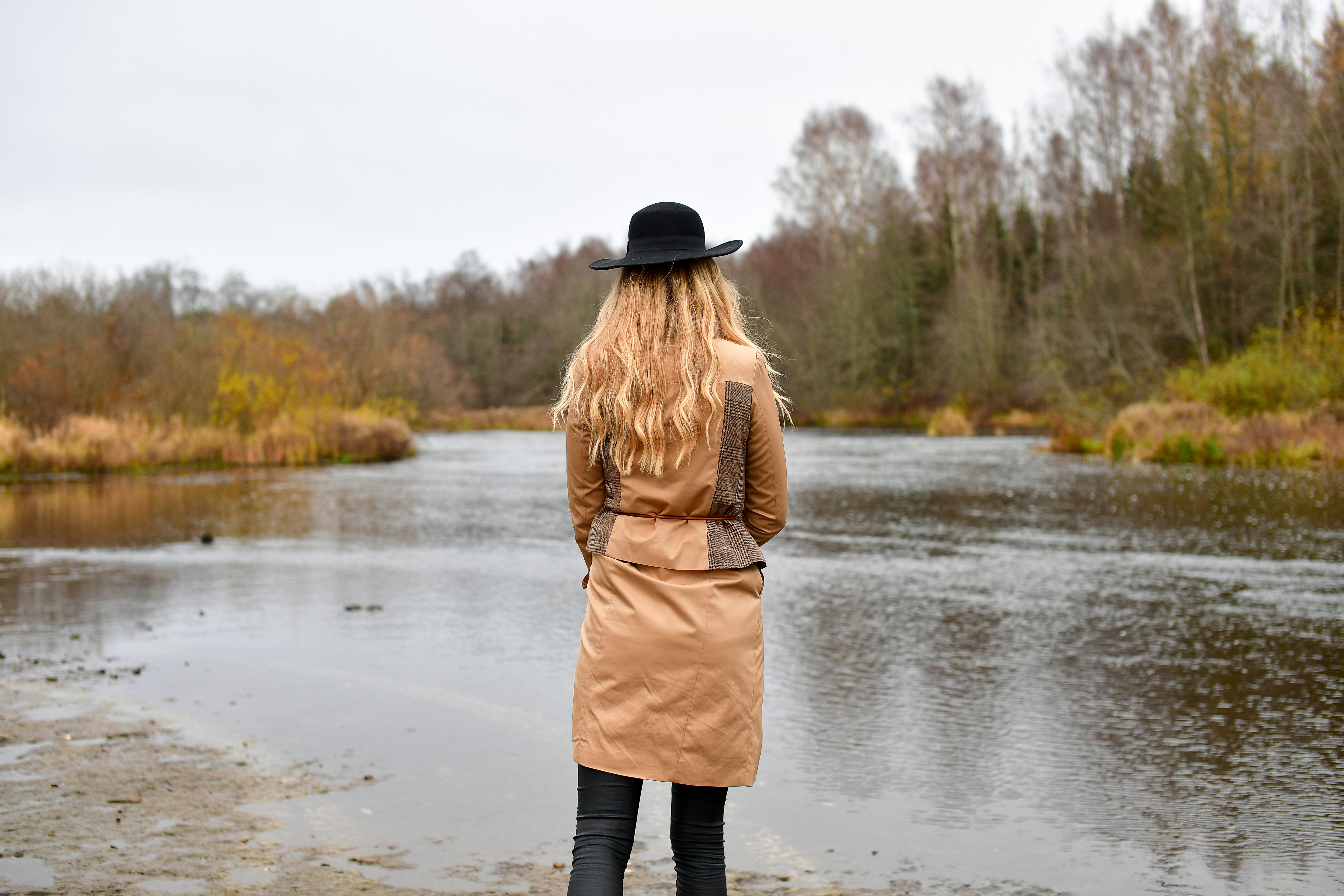Back view of a stylish woman in a trench coat and black hat walking towards a serene river in autumn.