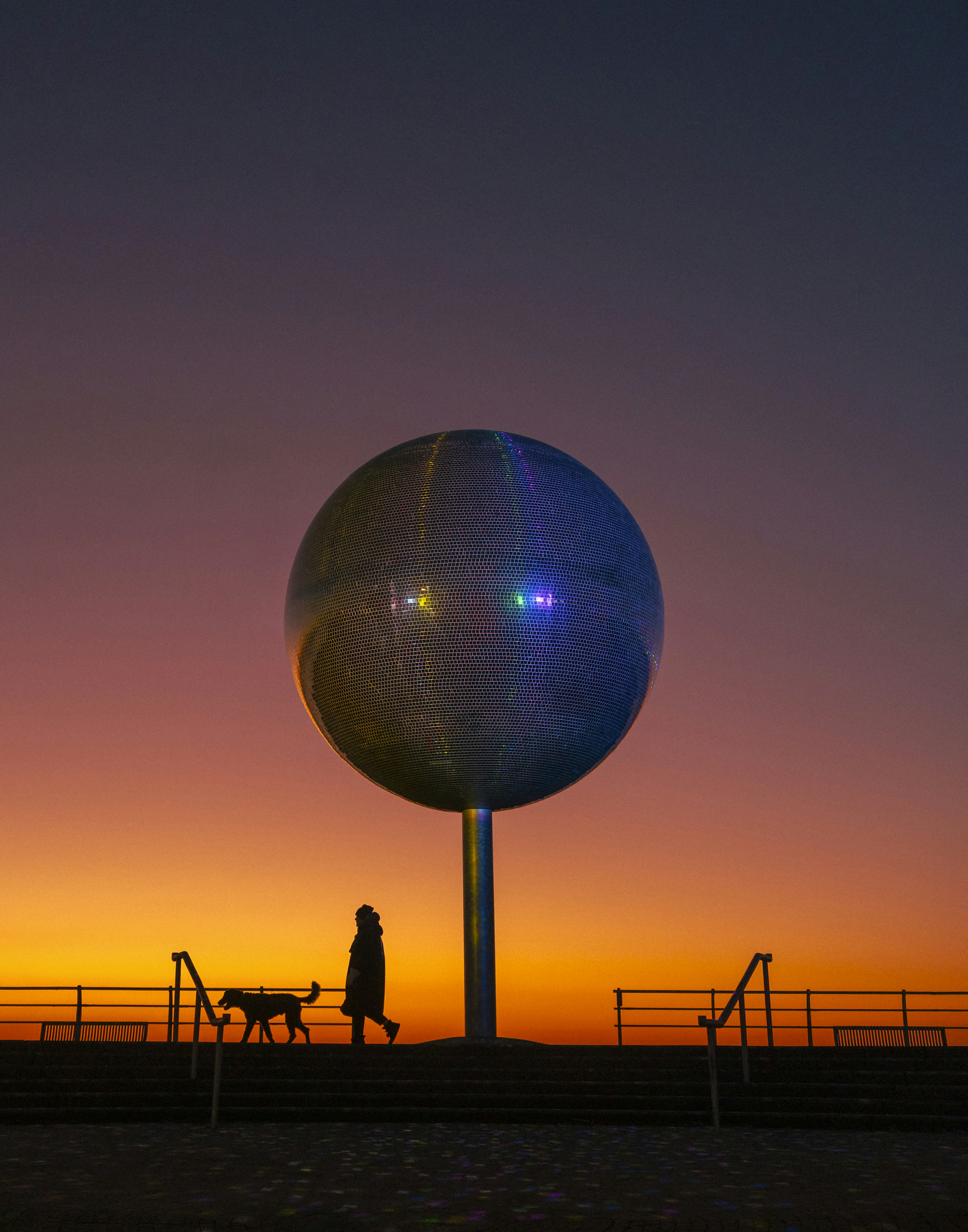 Person walking dog near large disco ball at sunset