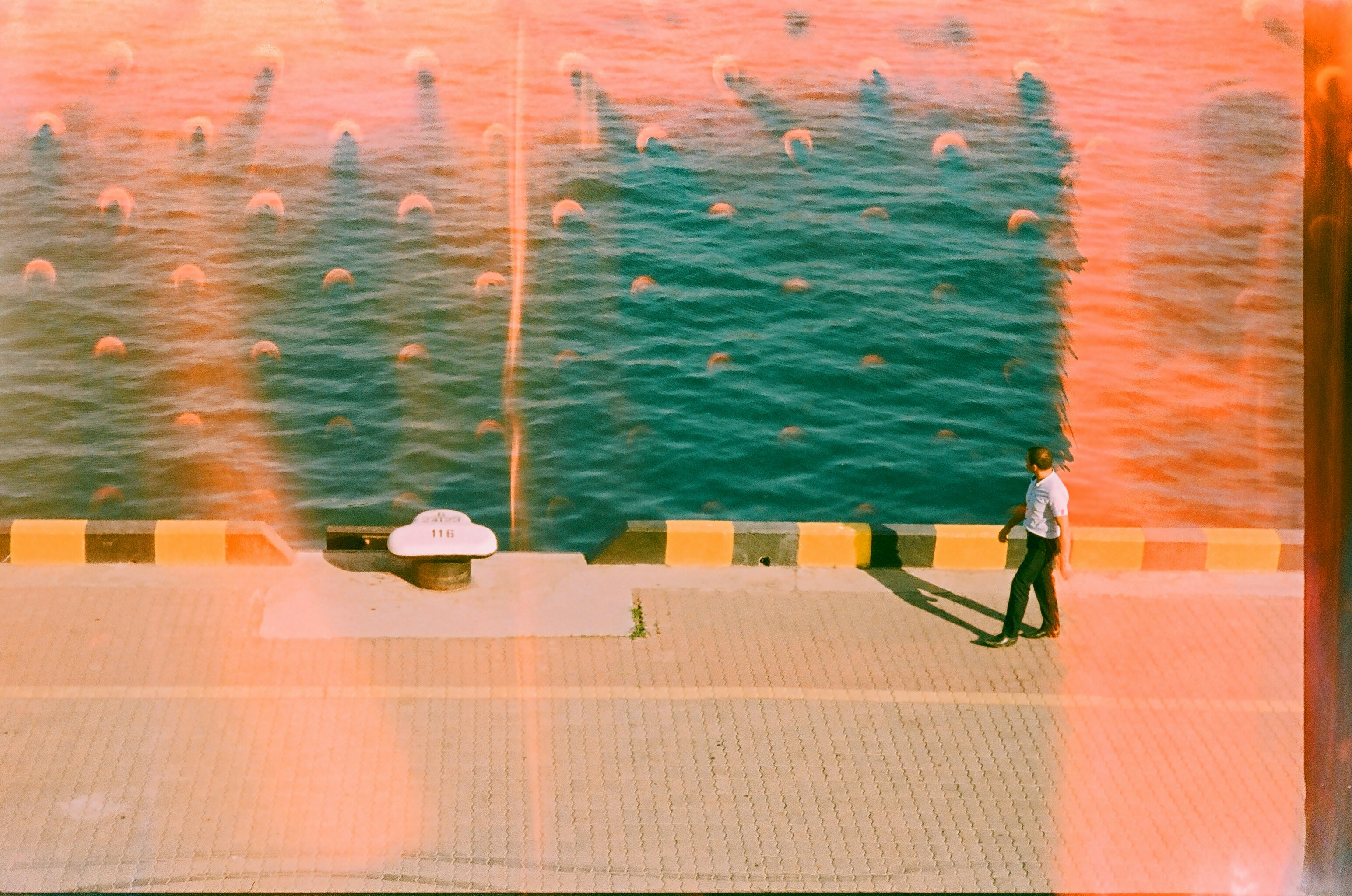Man walks along a sunny waterfront with teal water.