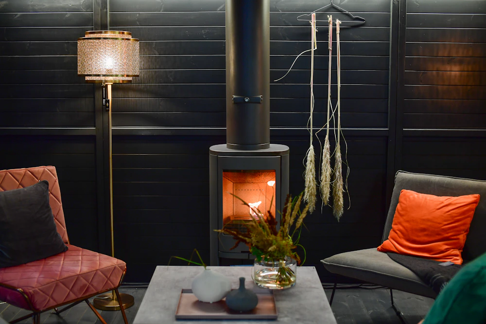 Two armchairs facing each other beside a lit wood stove, a lamp glowing against a dark wall