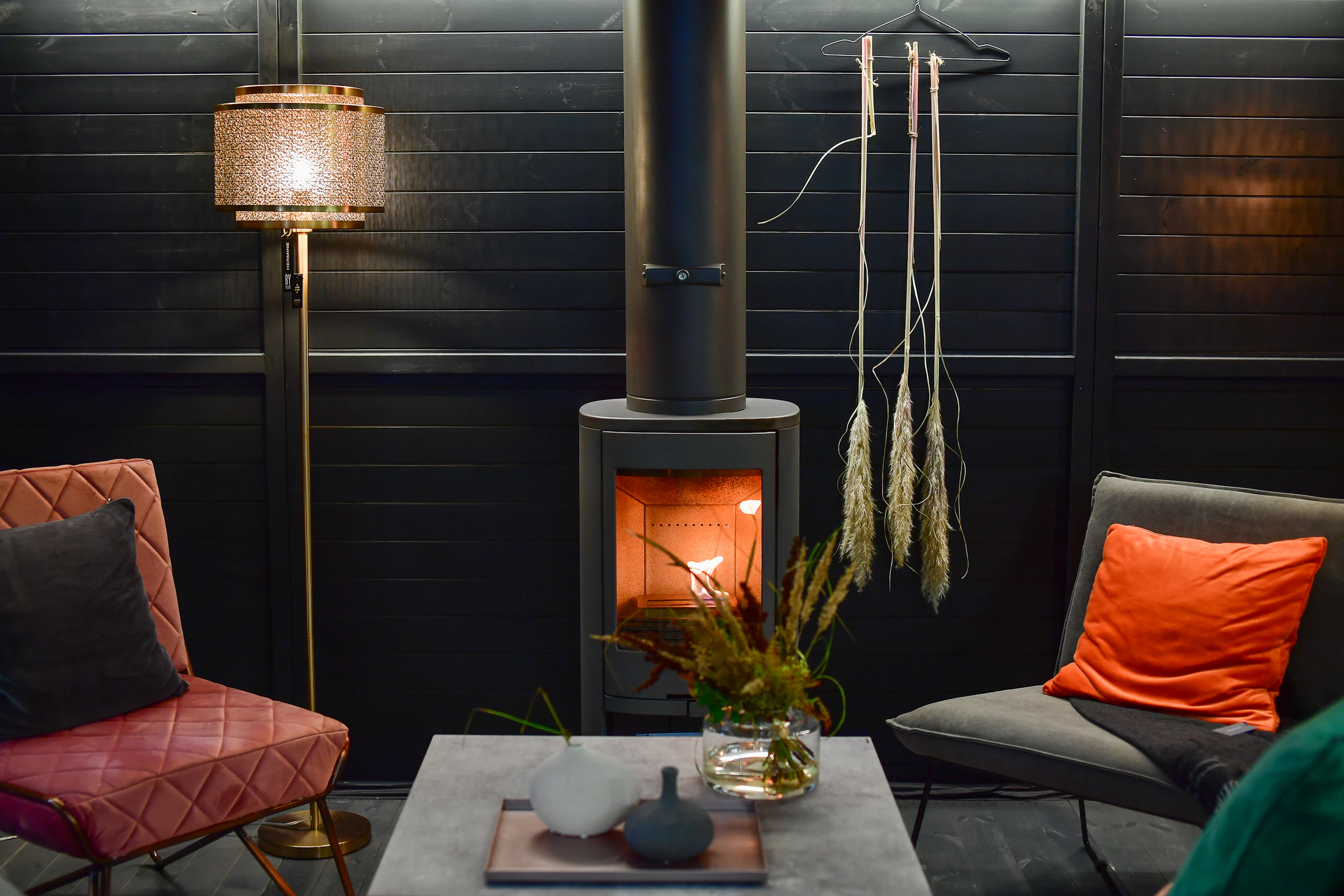 A dark living room with a lit wood stove, lounge chairs and warm lamplight on wood panelling