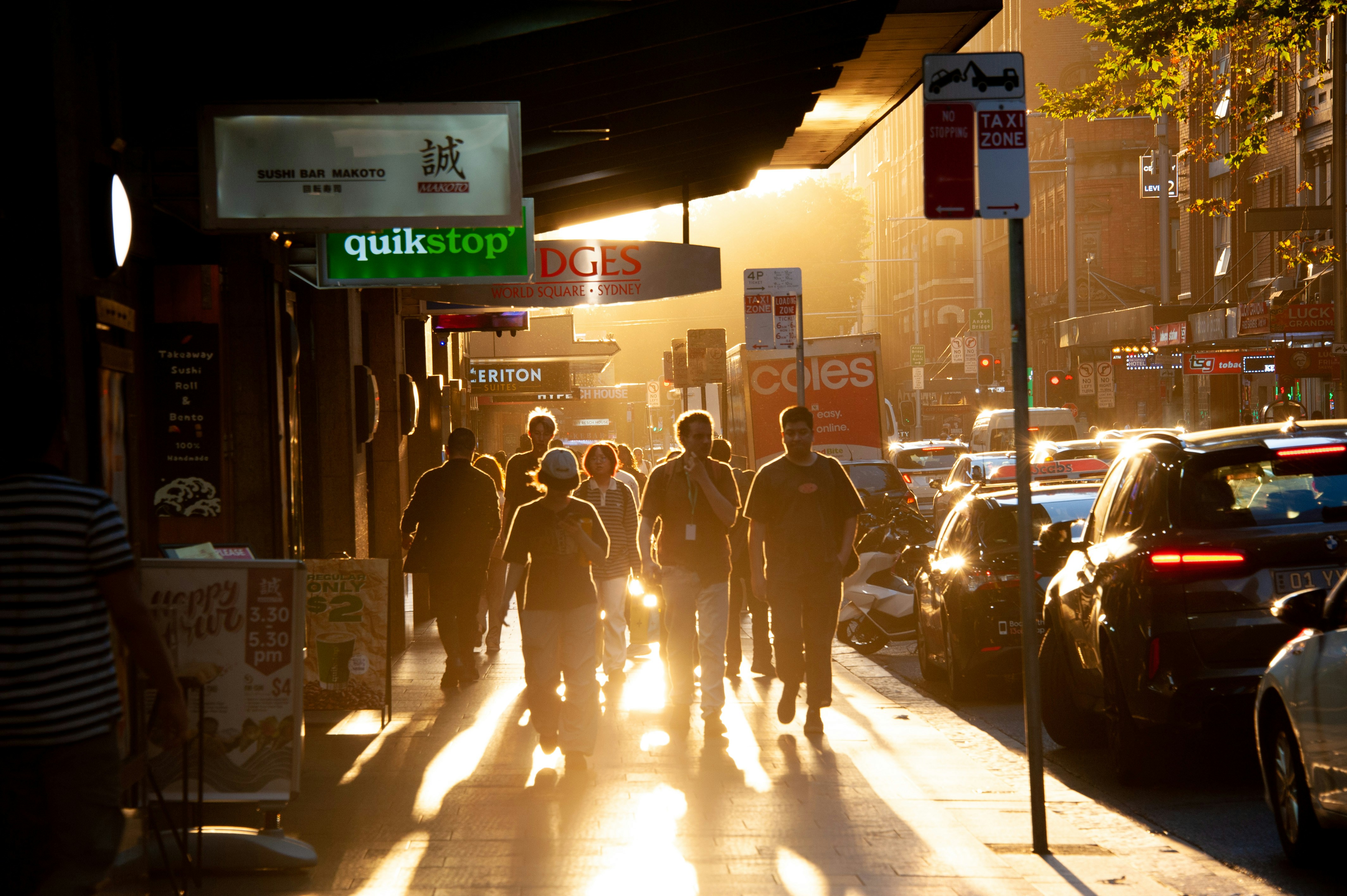 People walk down a sunlit city street with cars.