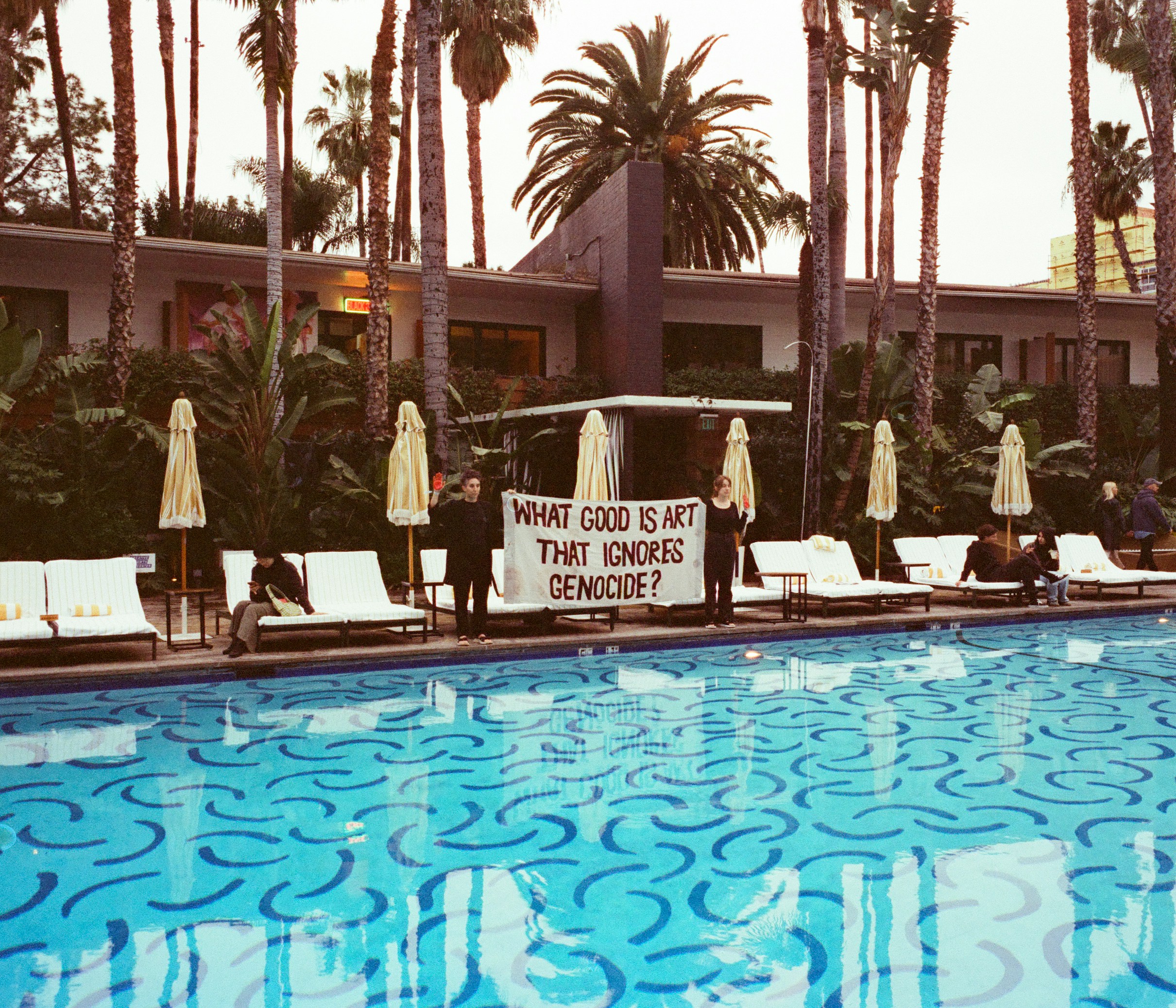 Protesters hold banner by swimming pool with palm trees.